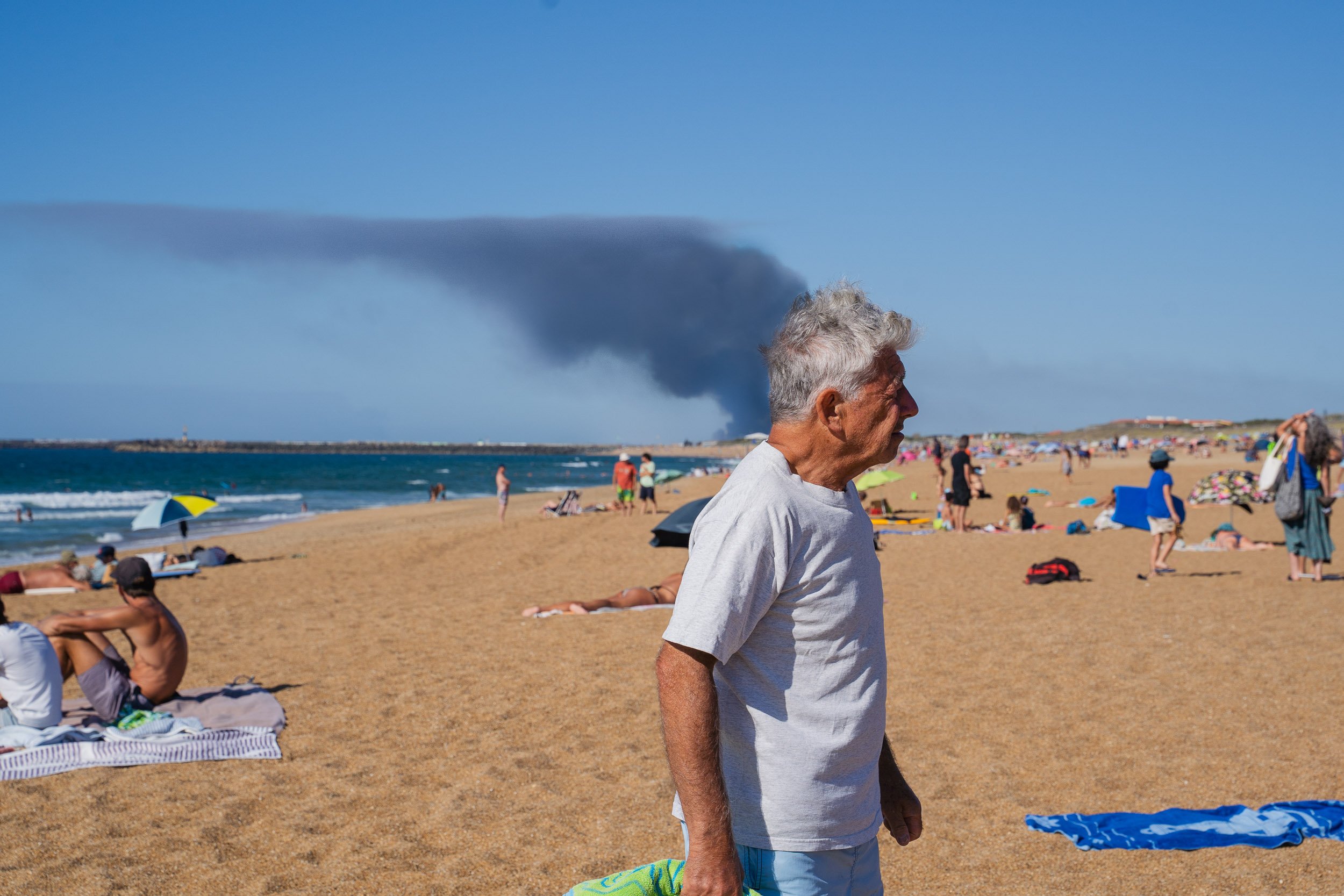 An elderly man walking on a crowded beach with people sunbathing and umbrellas, with a large smoke plume in the background.