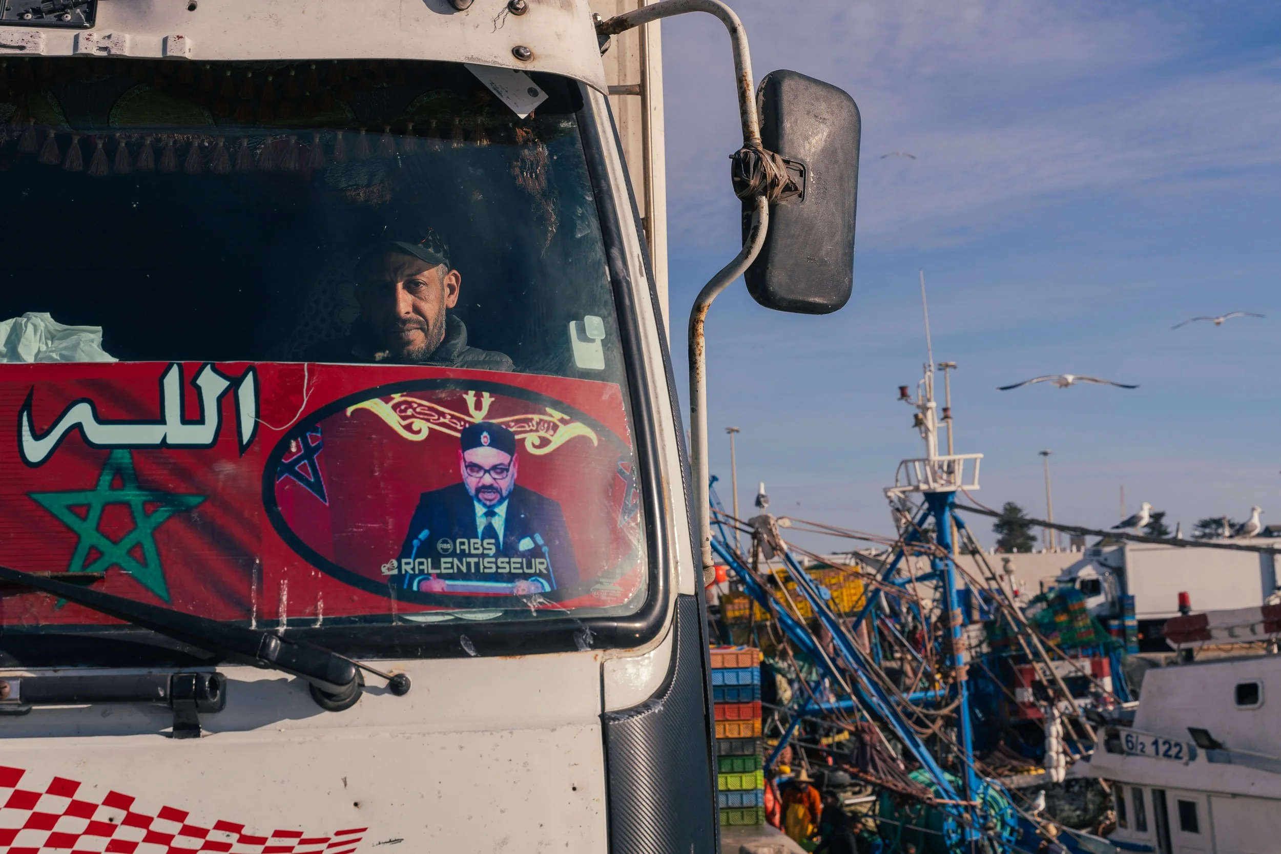 A man inside a large truck with a poster on the windshield, showing a man wearing glasses and a suit, with a Moroccan flag and Arabic text. Outside the truck, there are seagulls flying and a busy harbor with colorful fishing boats and equipment.