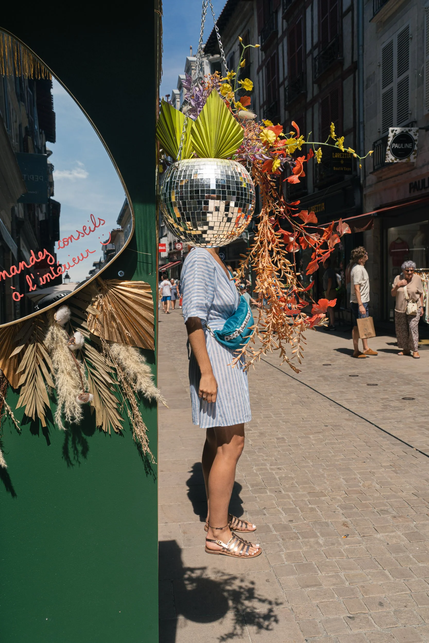 Person standing on a cobblestone street in a striped dress, wearing sandals and a blue fanny pack, with a reflective disco ball and colorful decorative plants hanging in front of them, capturing a lively urban scene.