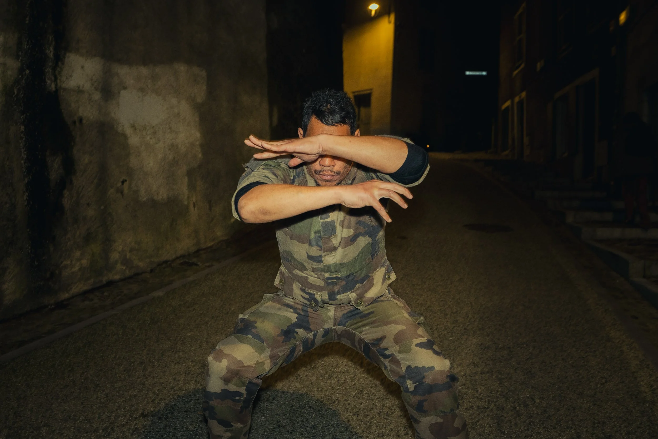 A man in camouflage military uniform performing a martial arts move in a dimly lit alley at night.