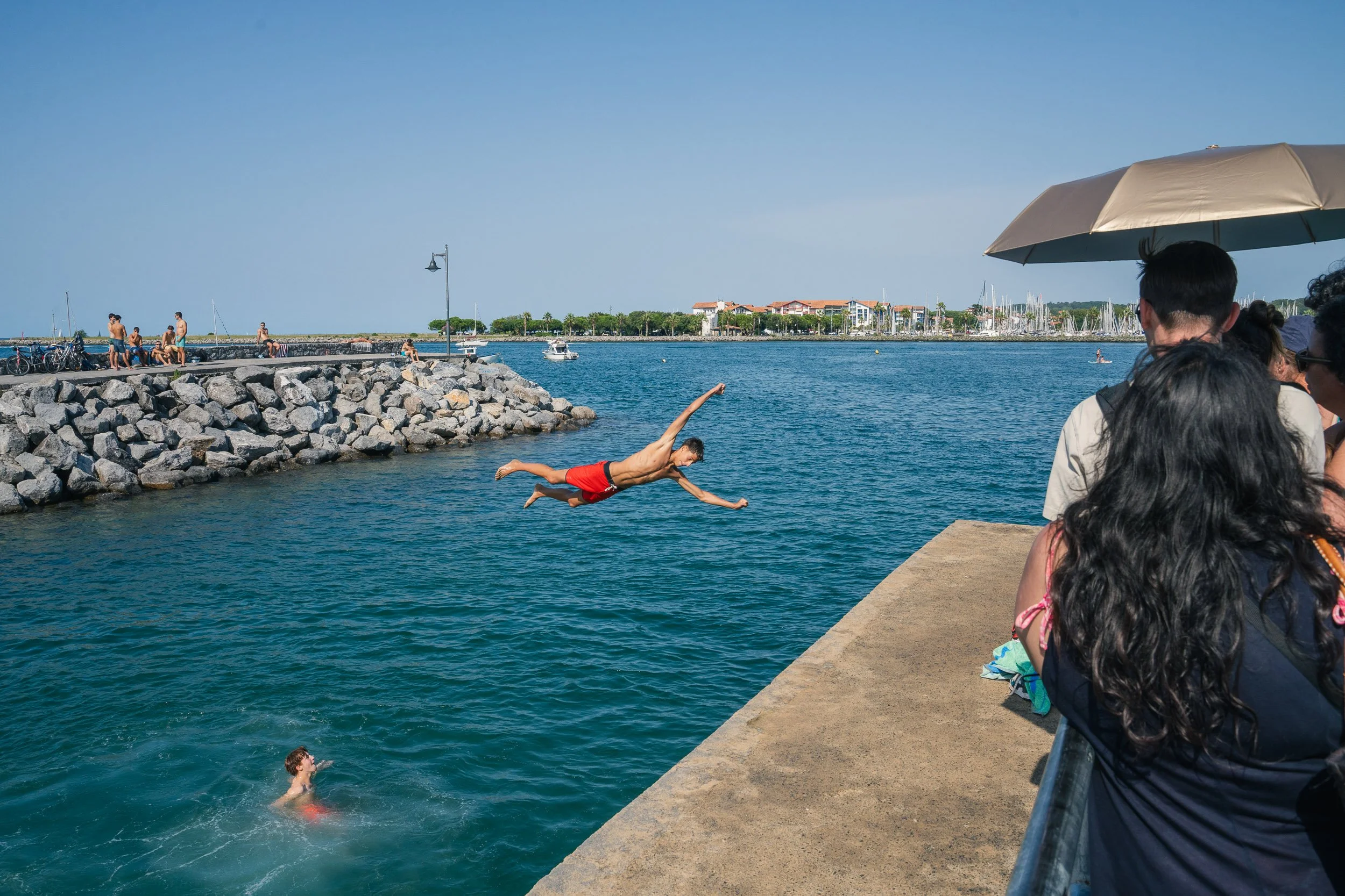 A man in red swimming trunks diving off a pier into a body of water, with people watching from the pier and swimmers in the water, and a harbor with boats and buildings in the background.