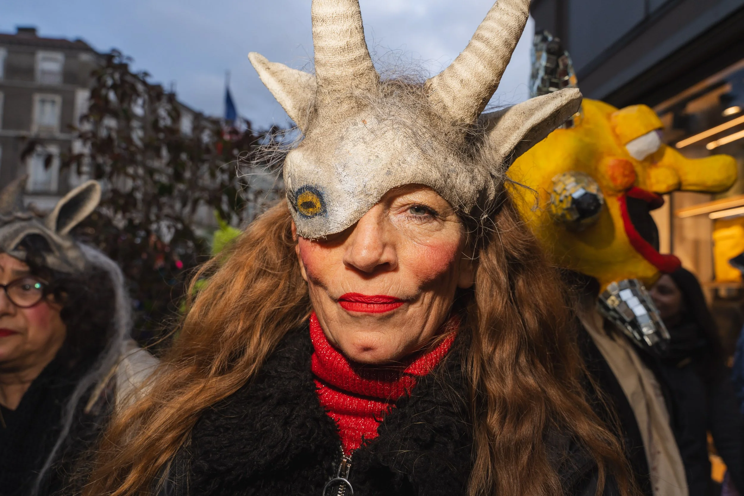 Woman wearing a large goat mask with long red hair, red lipstick, and a black jacket, with people in costumes visible in the background during an outdoor event.
