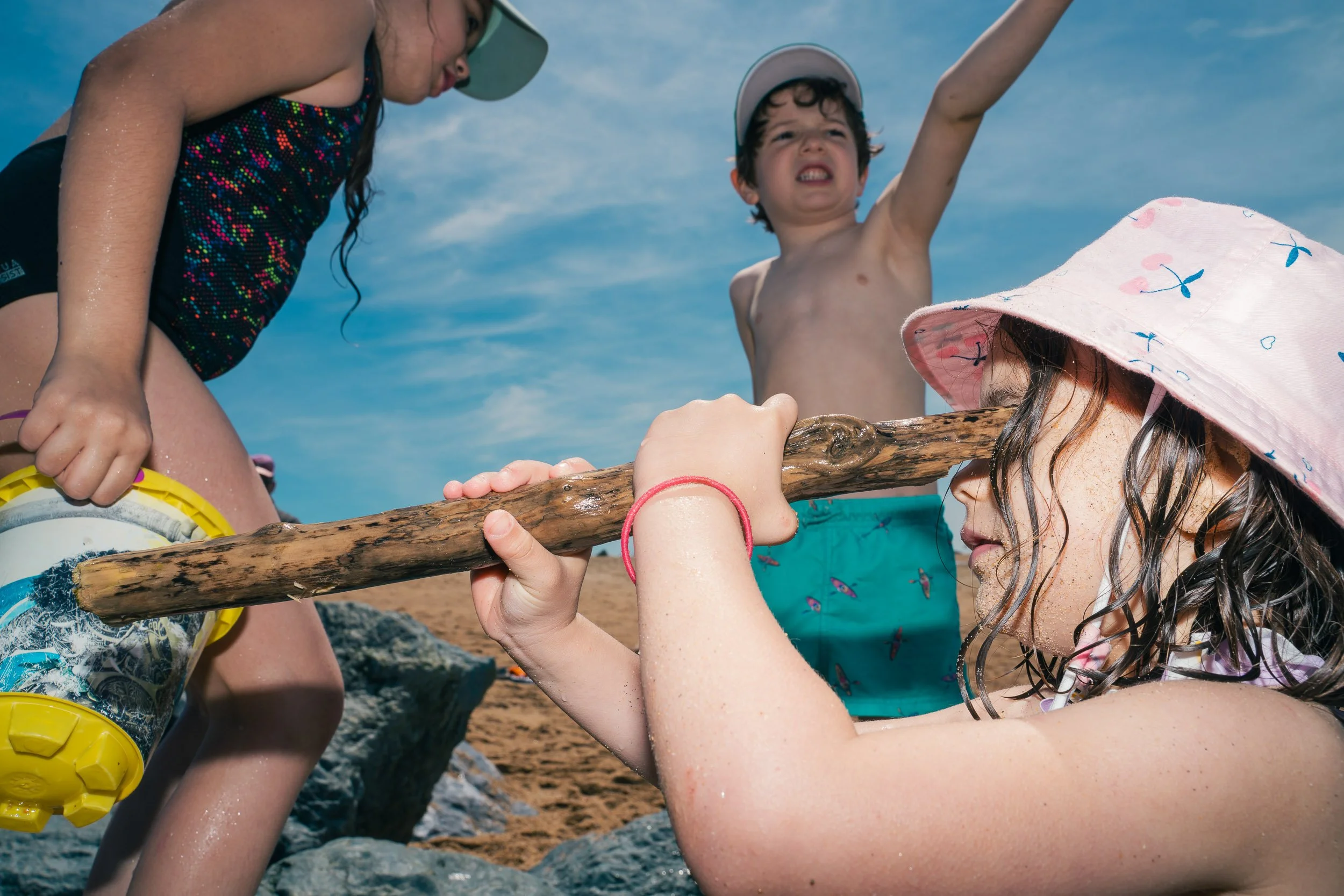 Children playing on the beach, with a girl holding a wooden stick and wearing a pink sunhat, a boy with a cap raising his arm, and another girl bending over, all under a blue sky.