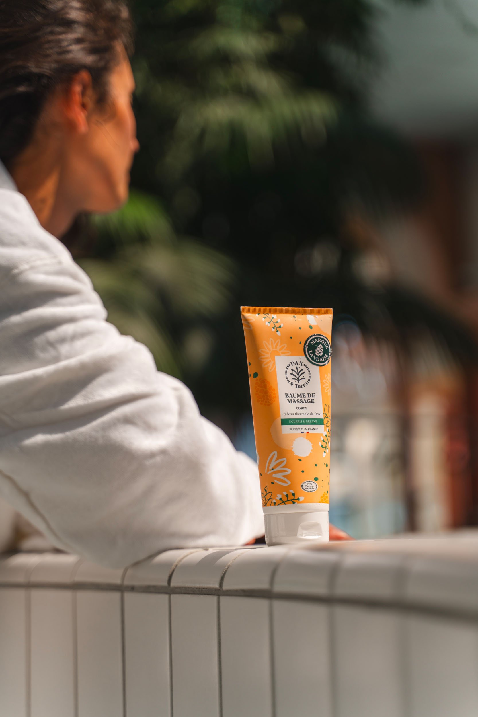 A person with short hair and a white shirt sits at a white tiled counter, holding a tube of massage balm with colorful floral design on the front.