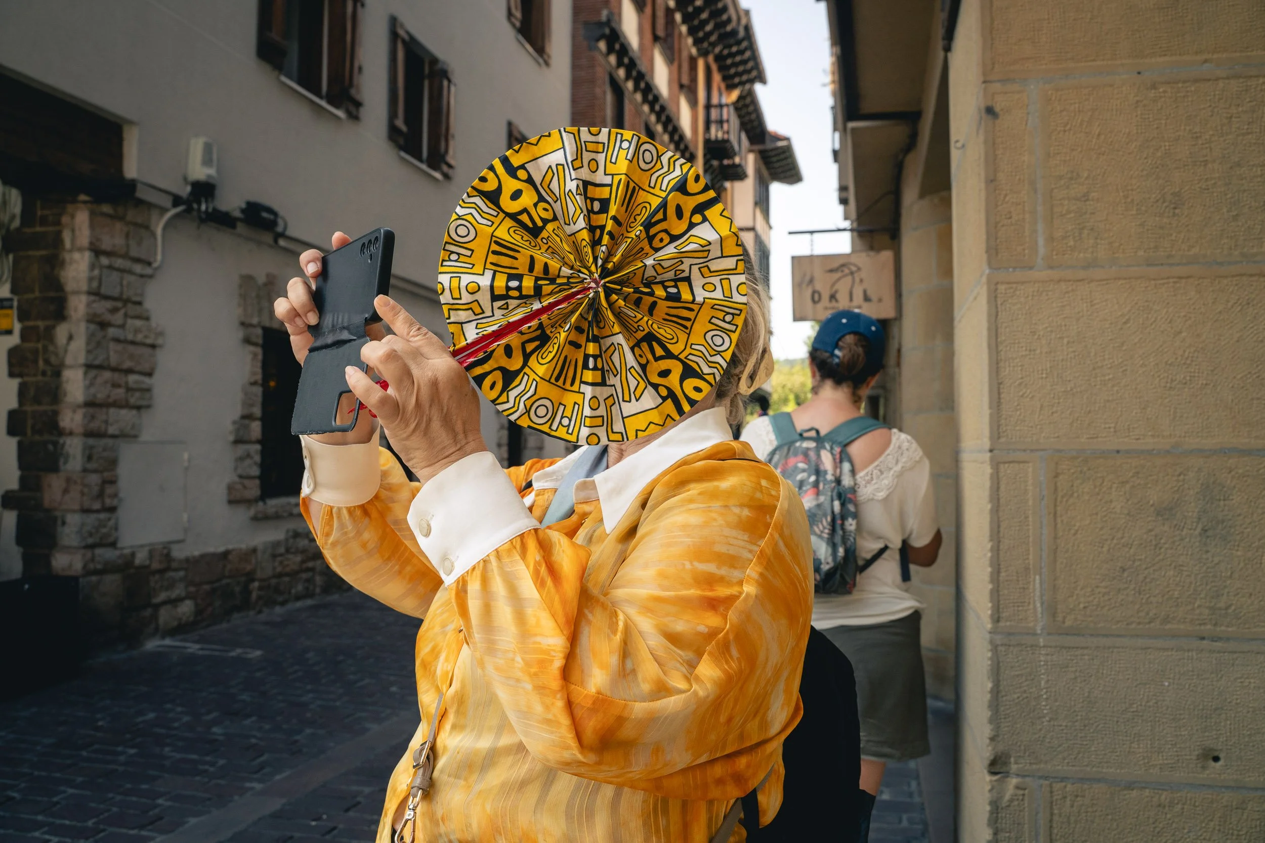 A woman wearing a yellow jacket is looking at her phone while holding a colorful umbrella with a yellow, black, and white abstract pattern. She is standing on a cobblestone street with two women in the background, one with a backpack and the other wi