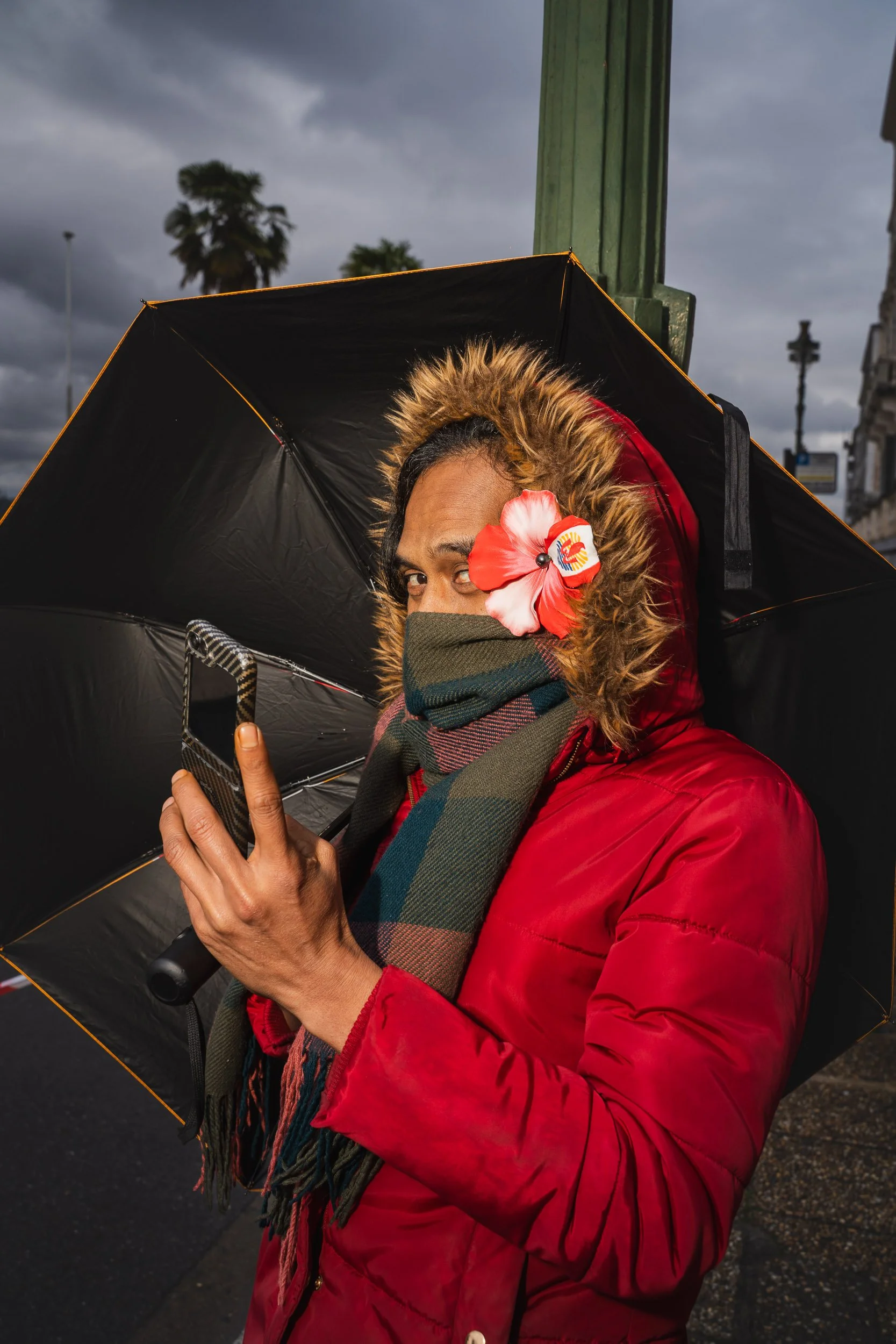 Woman wearing a red jacket, scarf, and a flower in her hair holding a smartphone under an umbrella and looking at the camera on a cloudy day.