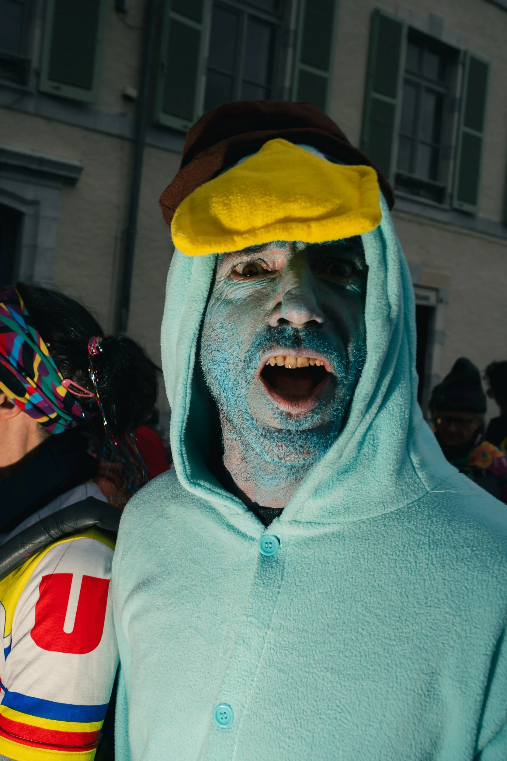 A person wearing a hoodie with a towel on their head, face painted with white and blue, and an expression of surprise or shock, at a street event or festival.