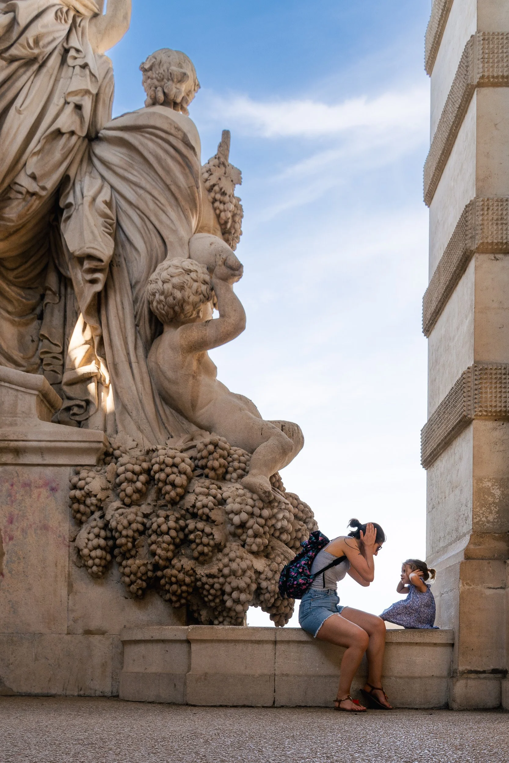 A woman and a young girl are sitting on a stone ledge, facing each other. The woman is taking a photo or video of the girl who is smiling and holding her hands up. They are outside near an ornate stone sculpture of cherubs and grapevine motifs agains