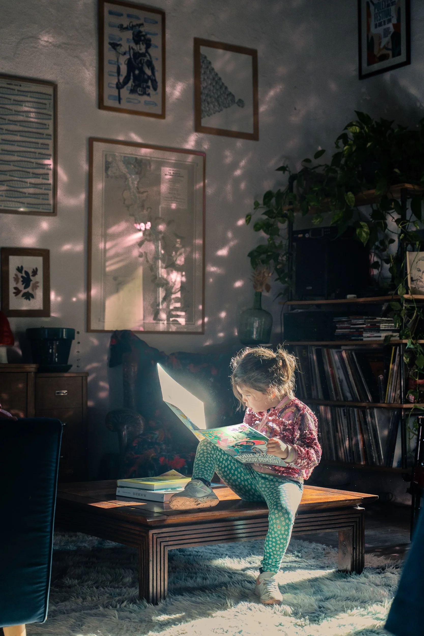 A young girl sitting cross-legged on a coffee table, reading a colorful magazine, with sunlight filtering through the window casting dappled shadows on the wall behind her.