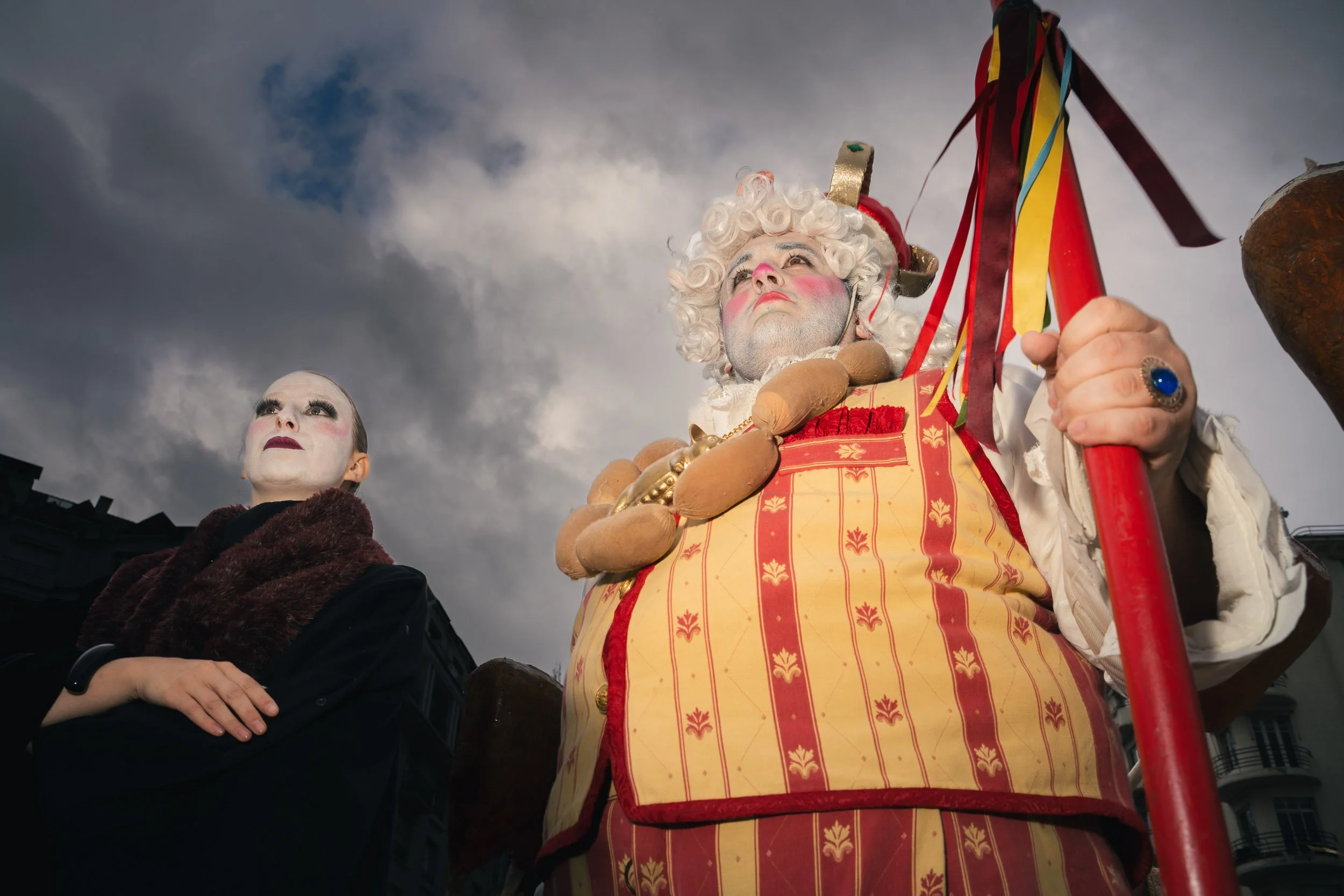 Two performers dressed as clowns or jesters are standing outdoors under a cloudy sky, with one holding a staff decorated with multicolored ribbons.