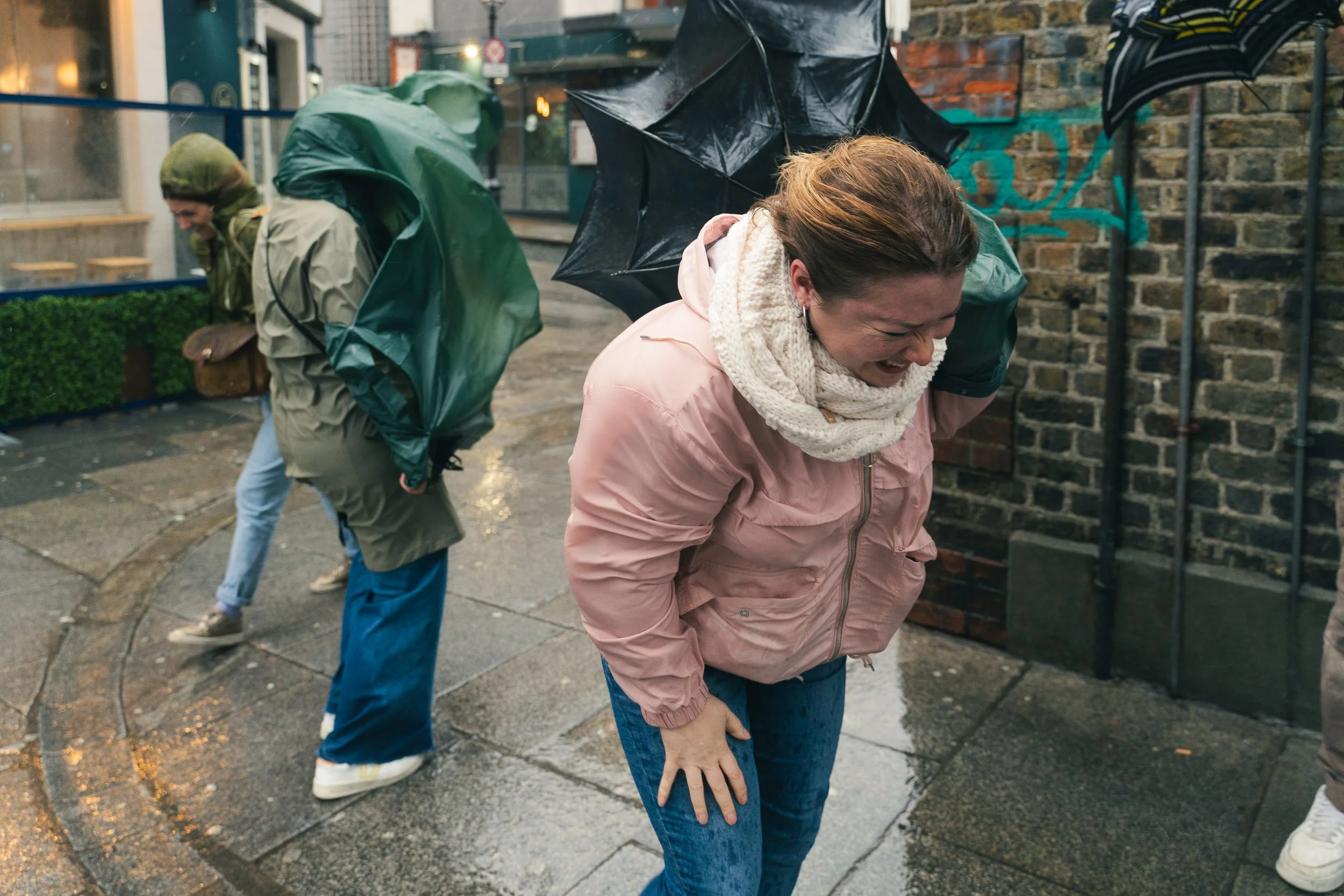 People standing on a wet sidewalk in the rain, with one woman in front wearing a pink jacket and scarf, holding her stomach and visibly upset, while others behind her appear to be searching for something or leaning over.