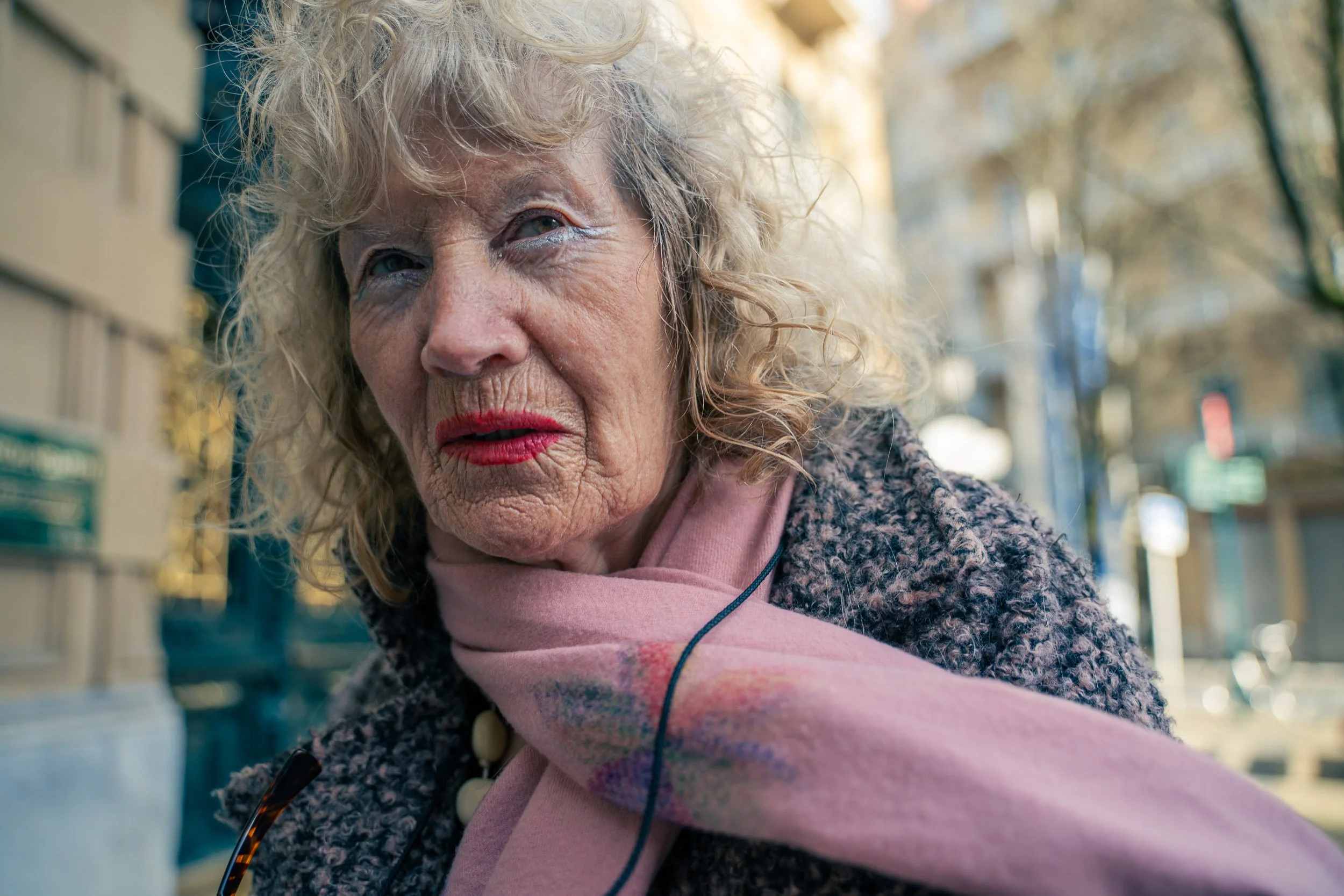 Close-up of an elderly woman with curly blonde hair, wearing a pink scarf and a textured coat, outdoors in an urban setting with trees and buildings in the background.