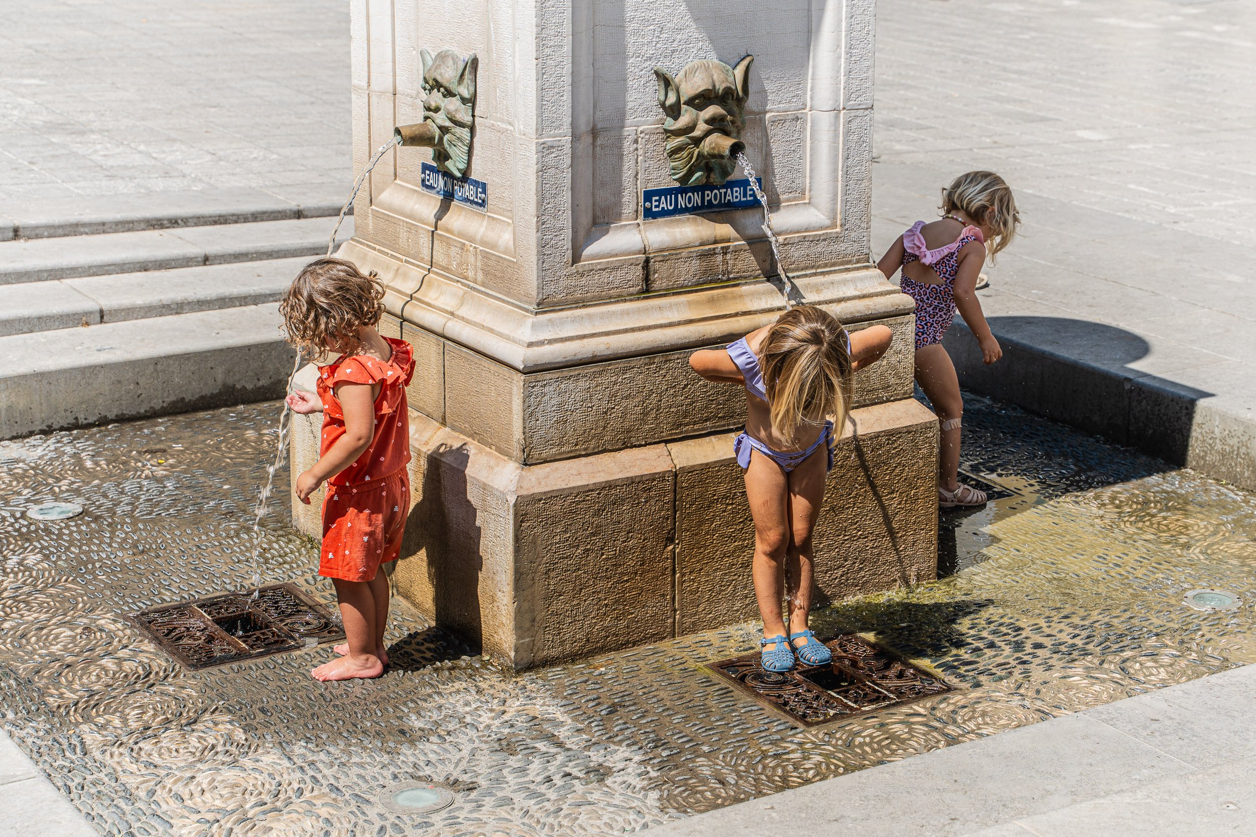 Three young girls playing in water fountains at a public fountain with water coming from a lion head spouts, on a sunny day.