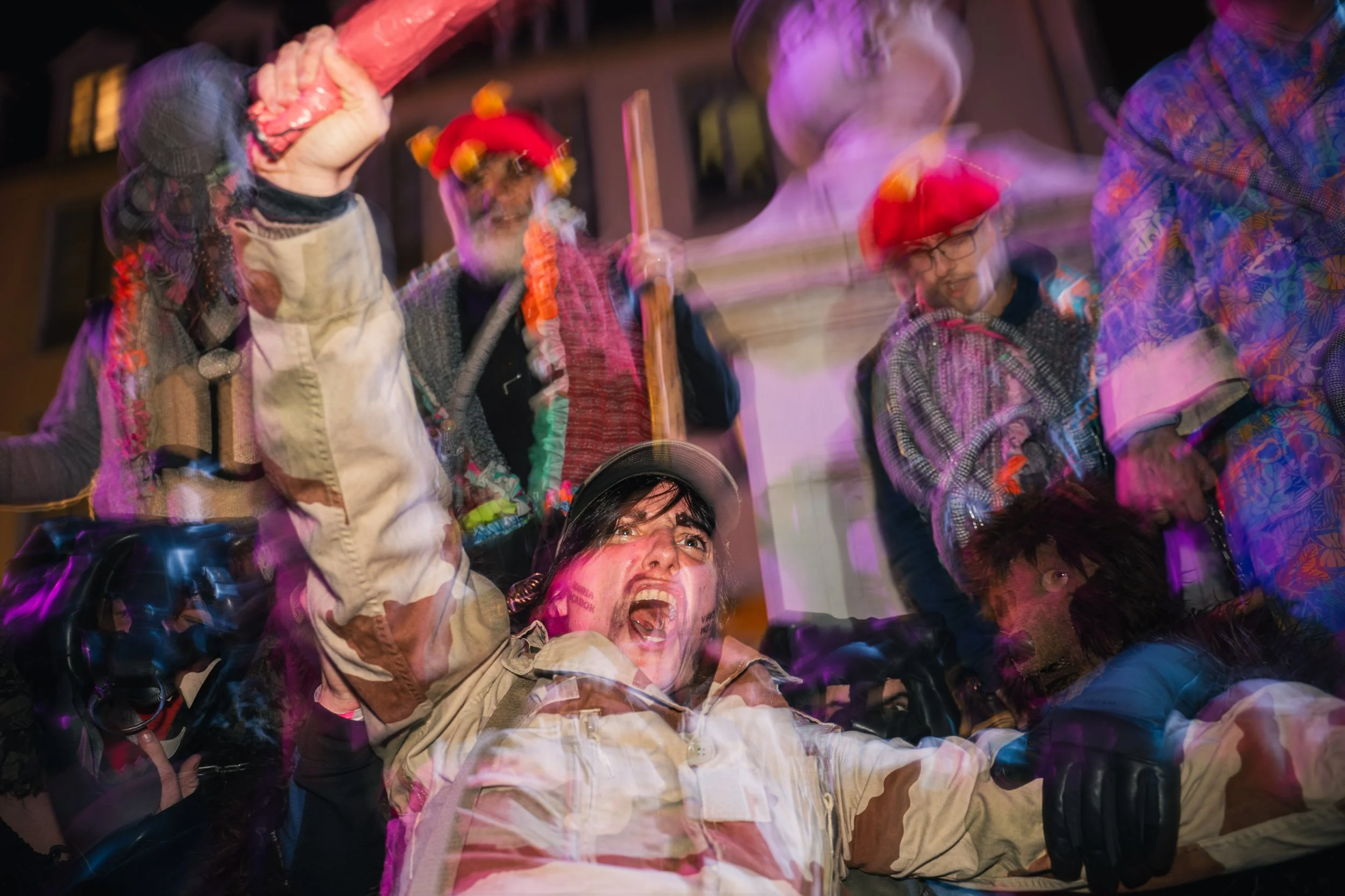 People celebrating at a night event, some wearing colorful costumes and wigs, with motion blur and vibrant lighting.