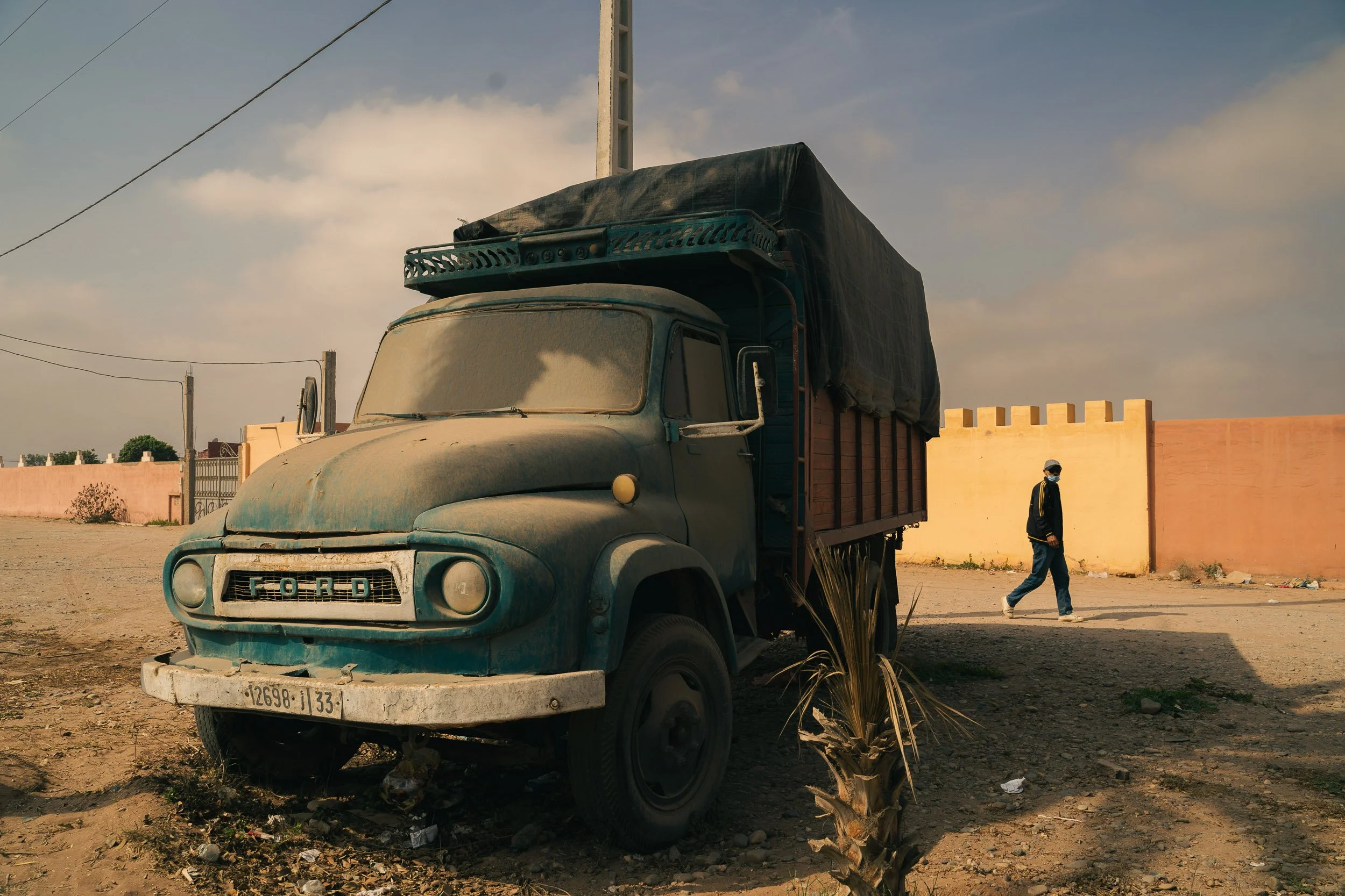 An old, dusty truck parked beside a dirt road, with a person walking by in the background, Morocco
