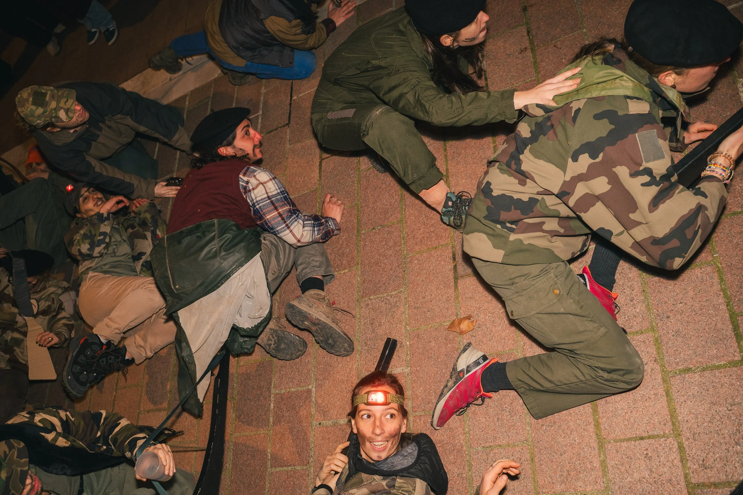 Group of young people lying on brick pavement, some wearing camouflage clothing and headlamps, appearing to be participating in an outdoor activity at night.