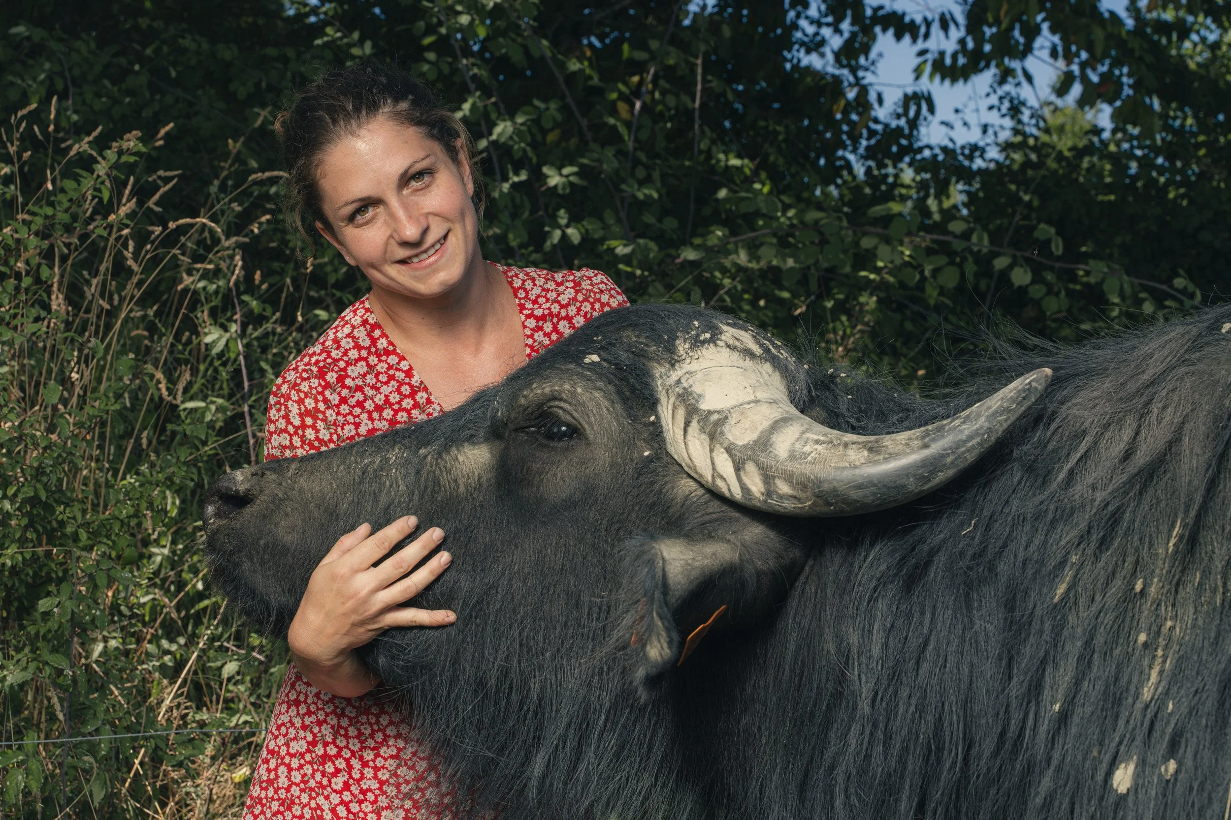 A woman in a red and white floral dress smiling while petting a large water buffalo outdoors with green foliage in the background.