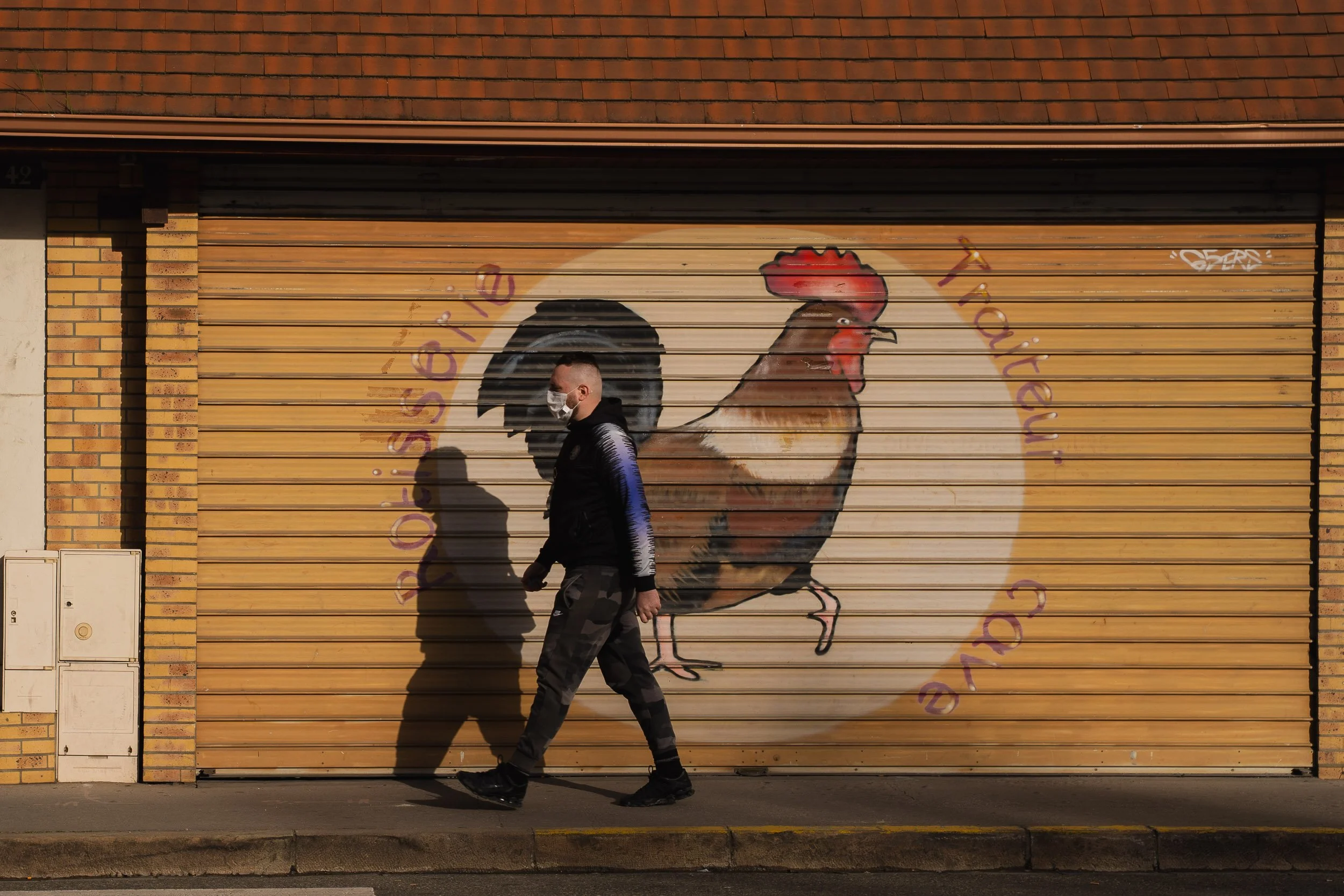 A man wearing a face mask and camouflage pants walking past a painted garage door featuring a rooster and a circle with the words "Ossigeno Ristorante" written around it.