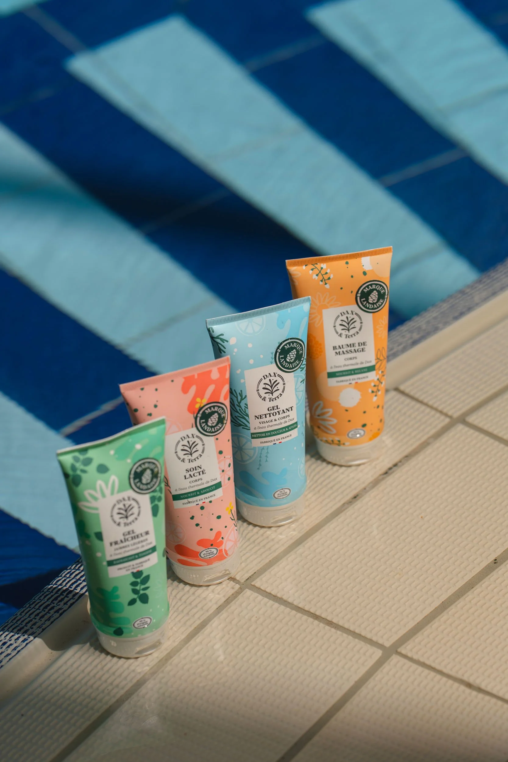 Four colorful tubes of skincare products placed on the edge of a swimming pool with blue and white striped tiles in the water.