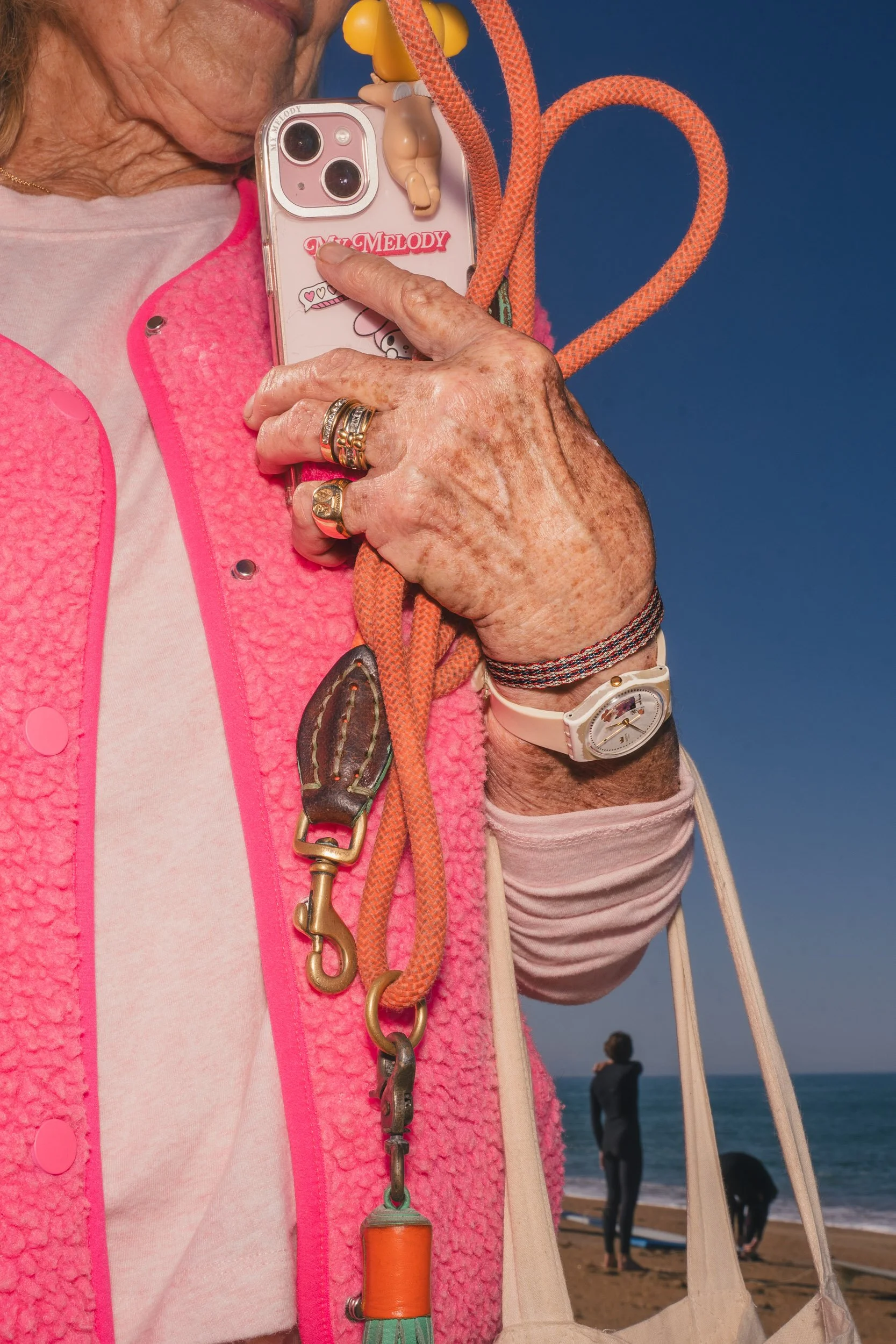 Close-up of an elderly woman's hand holding a smartphone with a pink case and a keychain, dressed in jewelry, with a pink fleece vest. The background shows a beach scene with a person looking at the ocean.