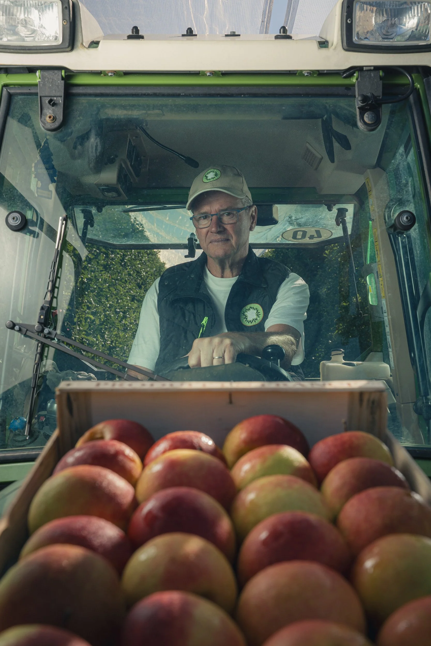 A man driving a green tractor, viewed from the front with apples in a crate in the foreground.