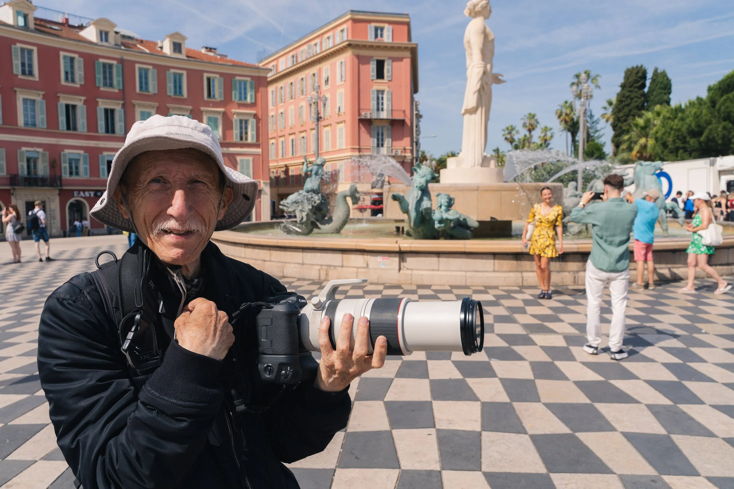 An elderly man in a black jacket and wide-brimmed hat holding a camera with a large white telephoto lens, standing in front of a fountain with sculptures and a statue in a paved plaza surrounded by colorful buildings with people taking photos and wal
