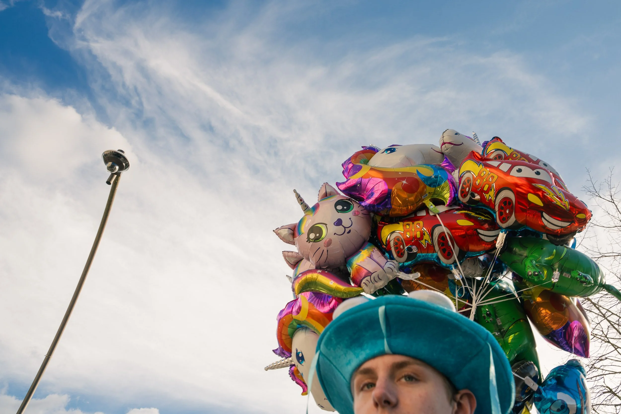 Close-up of a person's face, wearing a blue and white hat, with colorful balloons, including unicorn and car shapes, in the background against a partly cloudy sky.