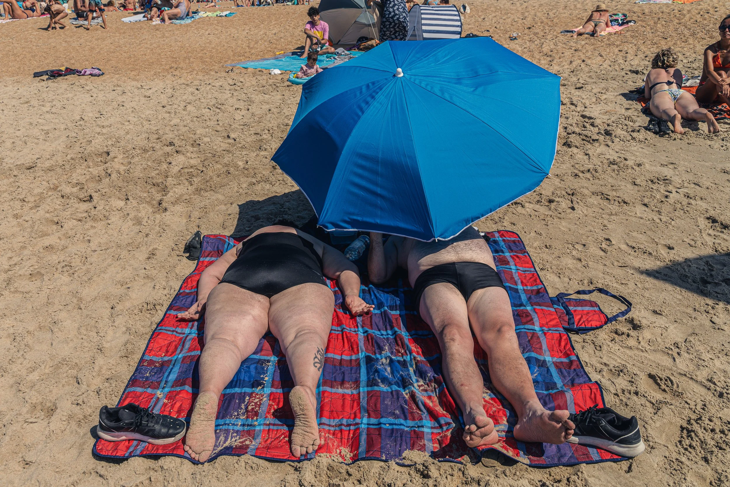Two people lying on a striped towel under a large blue umbrella on a crowded sandy beach, with other beachgoers in the background.