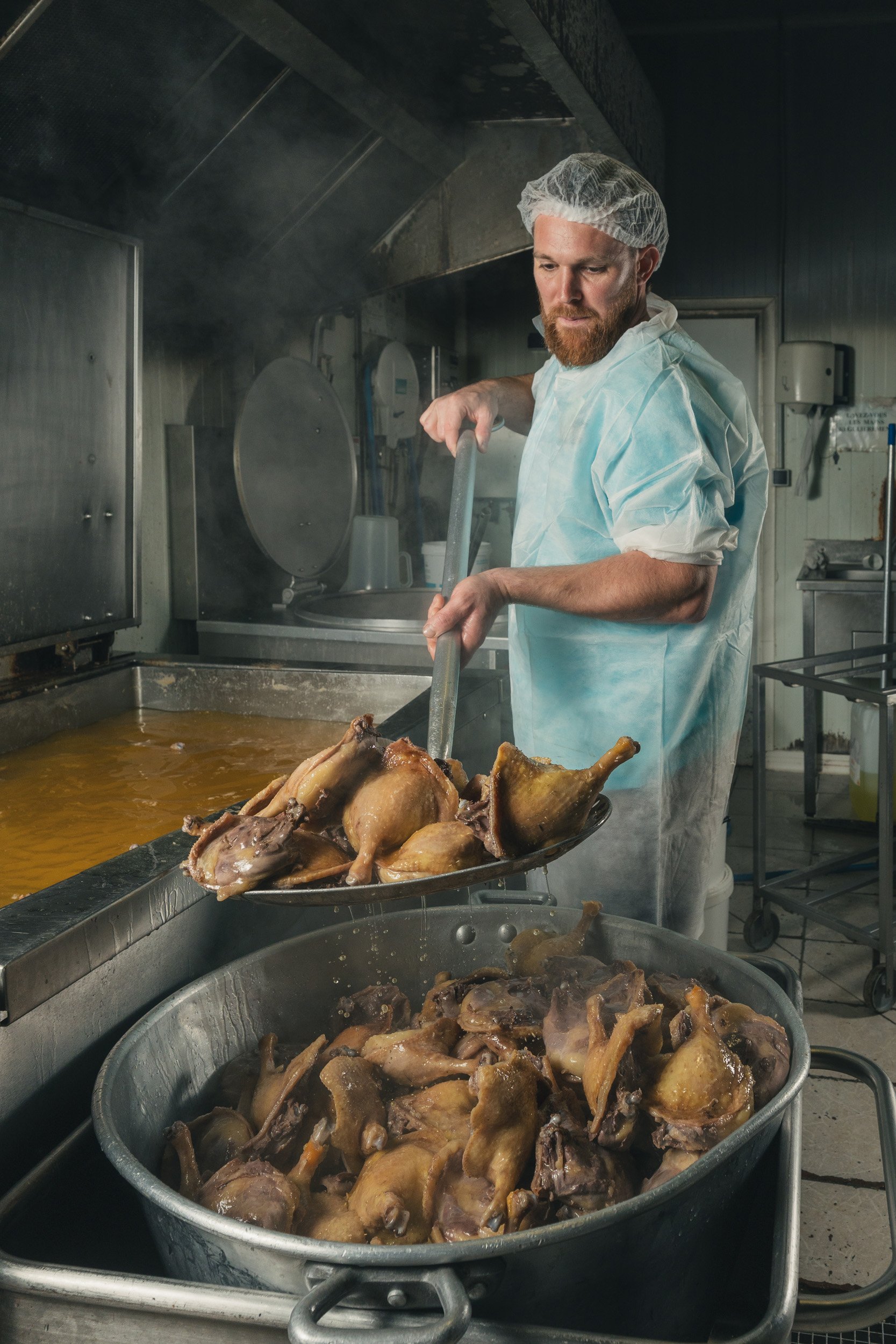 A man in a hair net and protective clothing is scooping cooked chicken into a large metal container in a commercial kitchen.