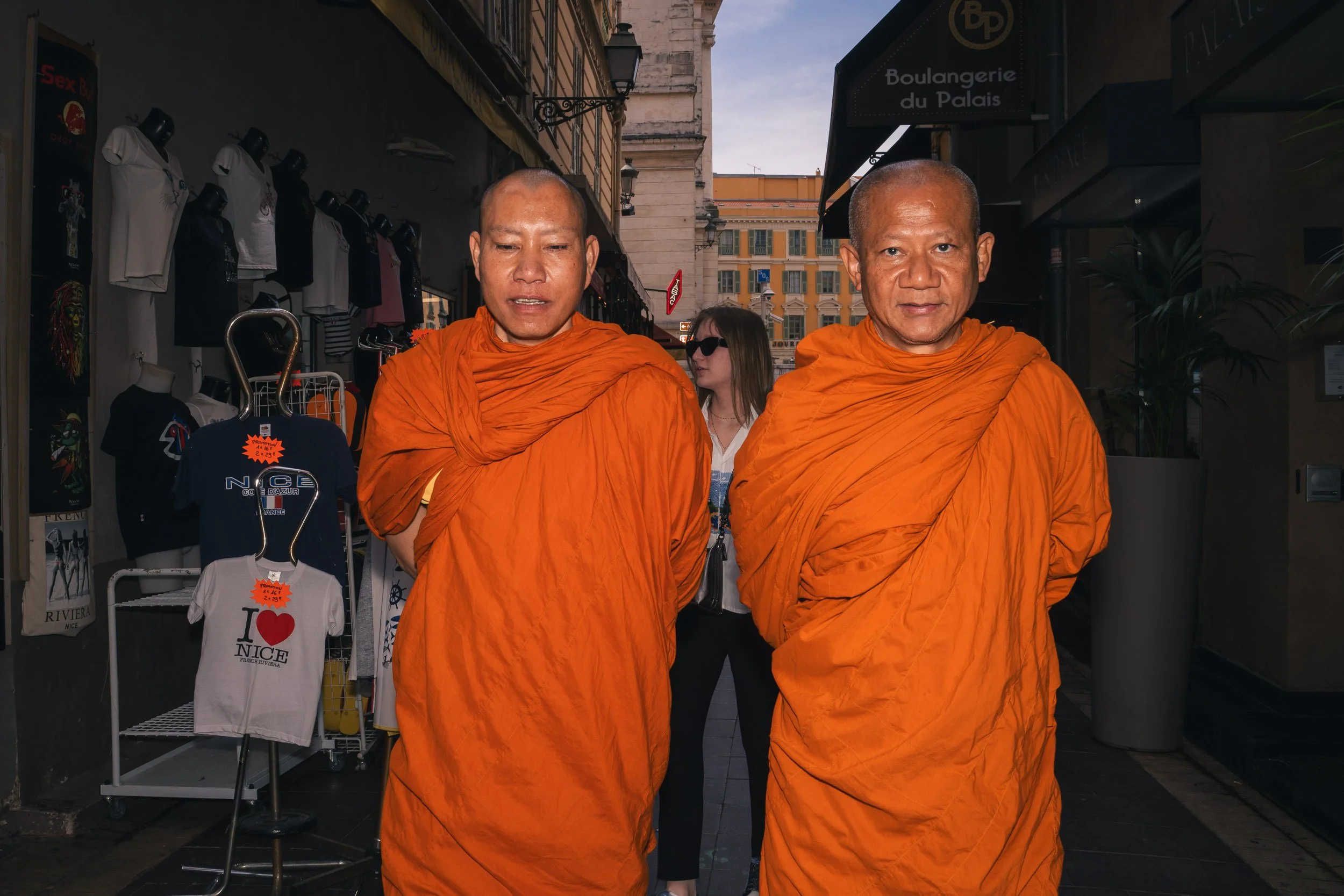 Two Buddhist monks dressed in orange robes walk down a street, with shops and a pedestrian in the background.