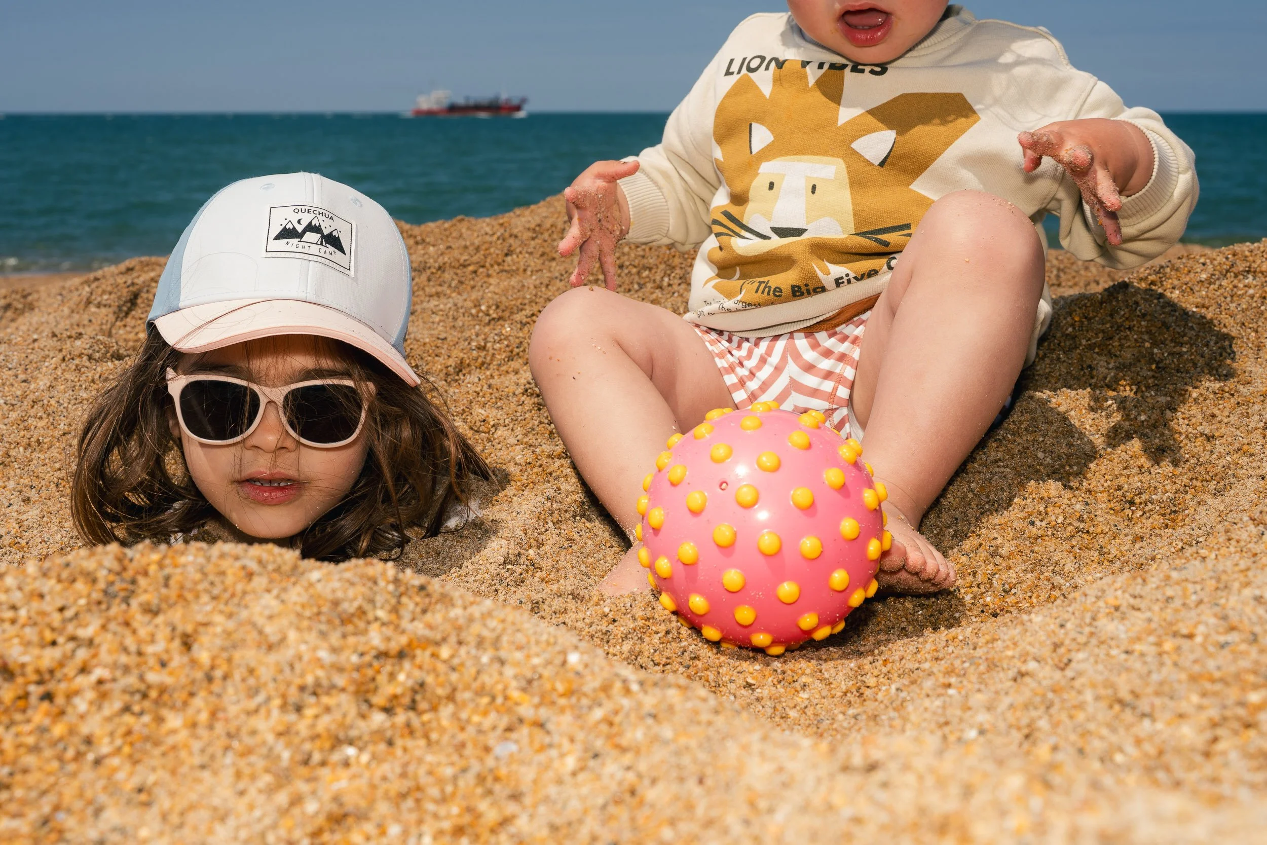 Two children playing on a sandy beach near the ocean; a girl with sunglasses and a cap is partially buried in the sand, while a boy with a lion graphic shirt is sitting next to a colorful spiky ball.