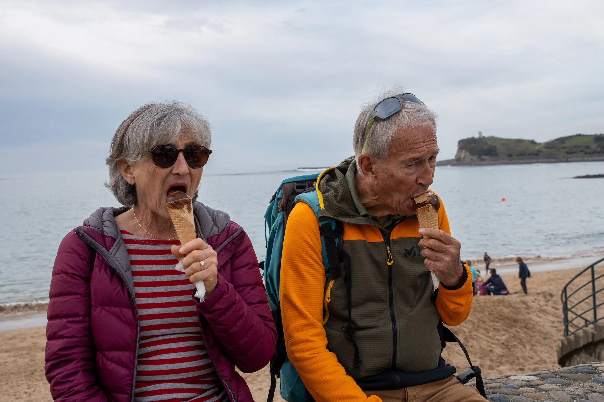 An elderly man and woman eating ice cream cones on a beach, with the ocean and cliffs in the background.
