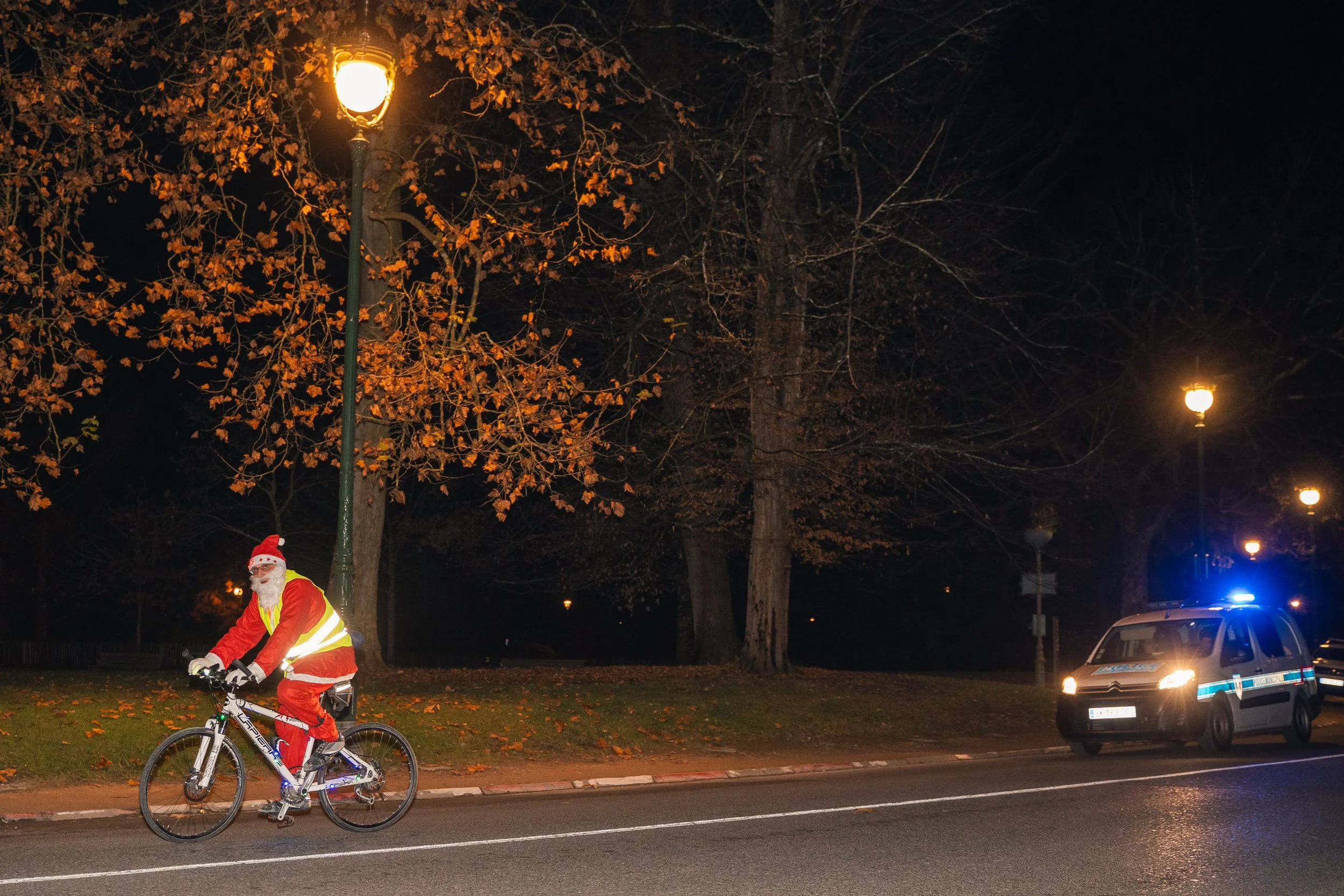 A person dressed as Santa Claus riding a bicycle at night with a police car parked nearby, illuminated by streetlights and surrounded by trees with autumn leaves.