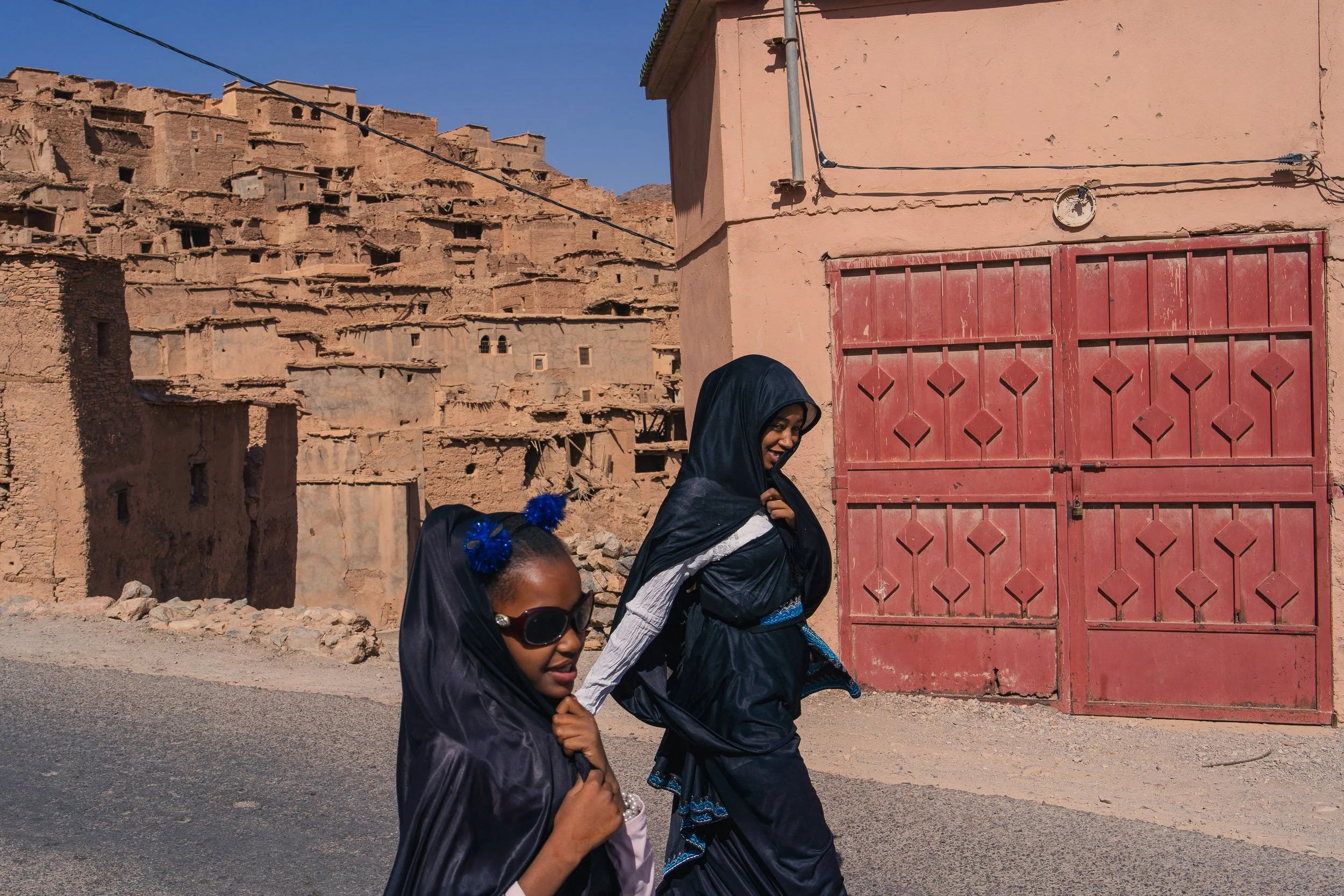 Two women walking on a dirt road in front of a pink building with a red door and an ancient mud-brick city in the background.