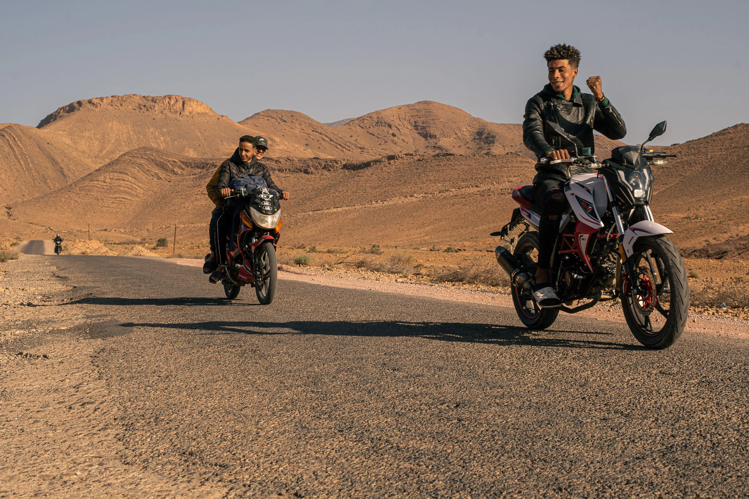 Two young men riding motorcycles on a desert road with rocky hills in the background, one smiling and holding a fist up, the other riding behind.