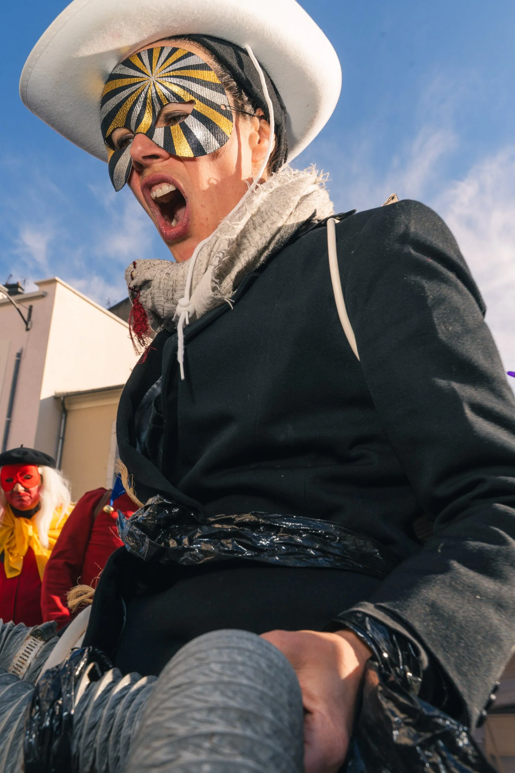 A person wearing a large white hat, black and gold patterned mask, and black jacket, shouting with an open mouth during a street event. Another person in the background has a red mask and yellow shirt.