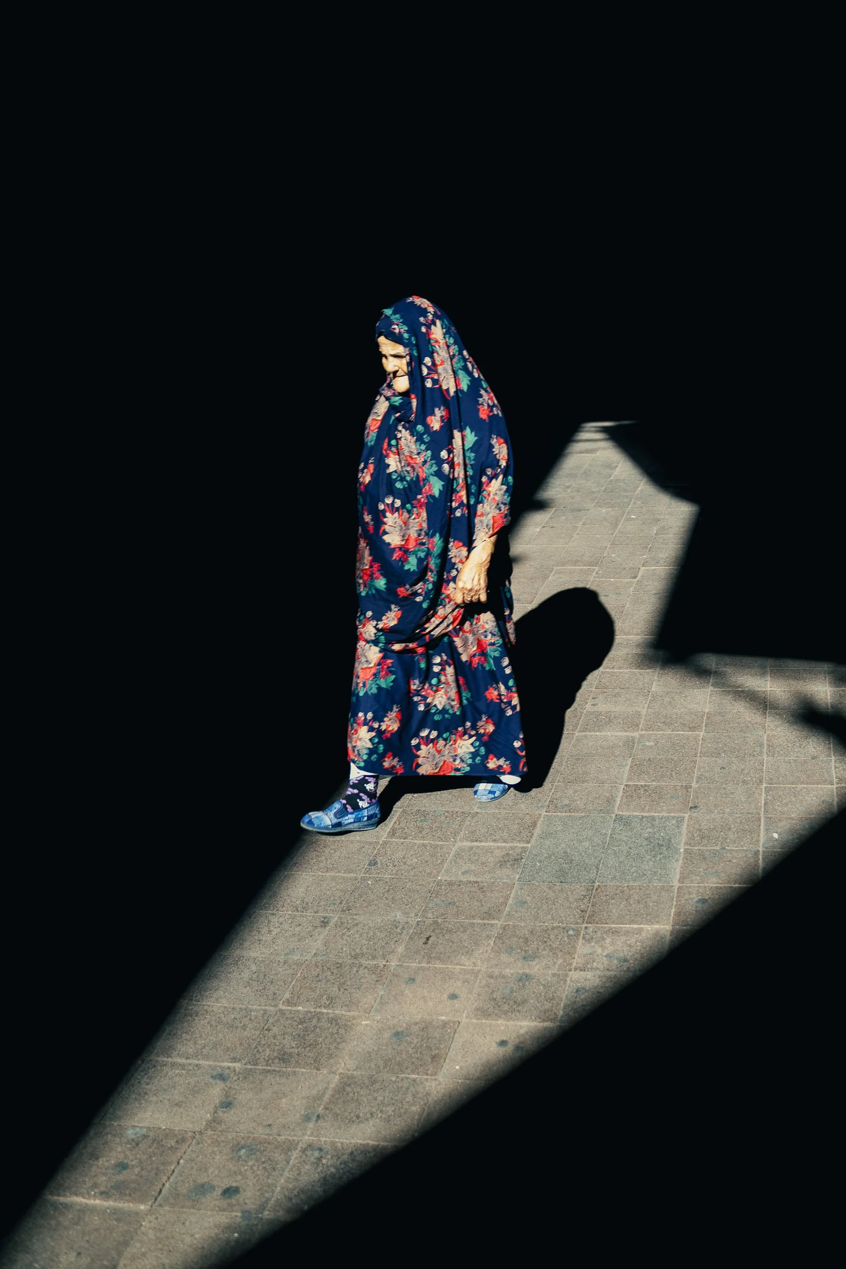 A woman wearing a floral patterned dress and headscarf walking on a sidewalk, with deep shadows cast around her.