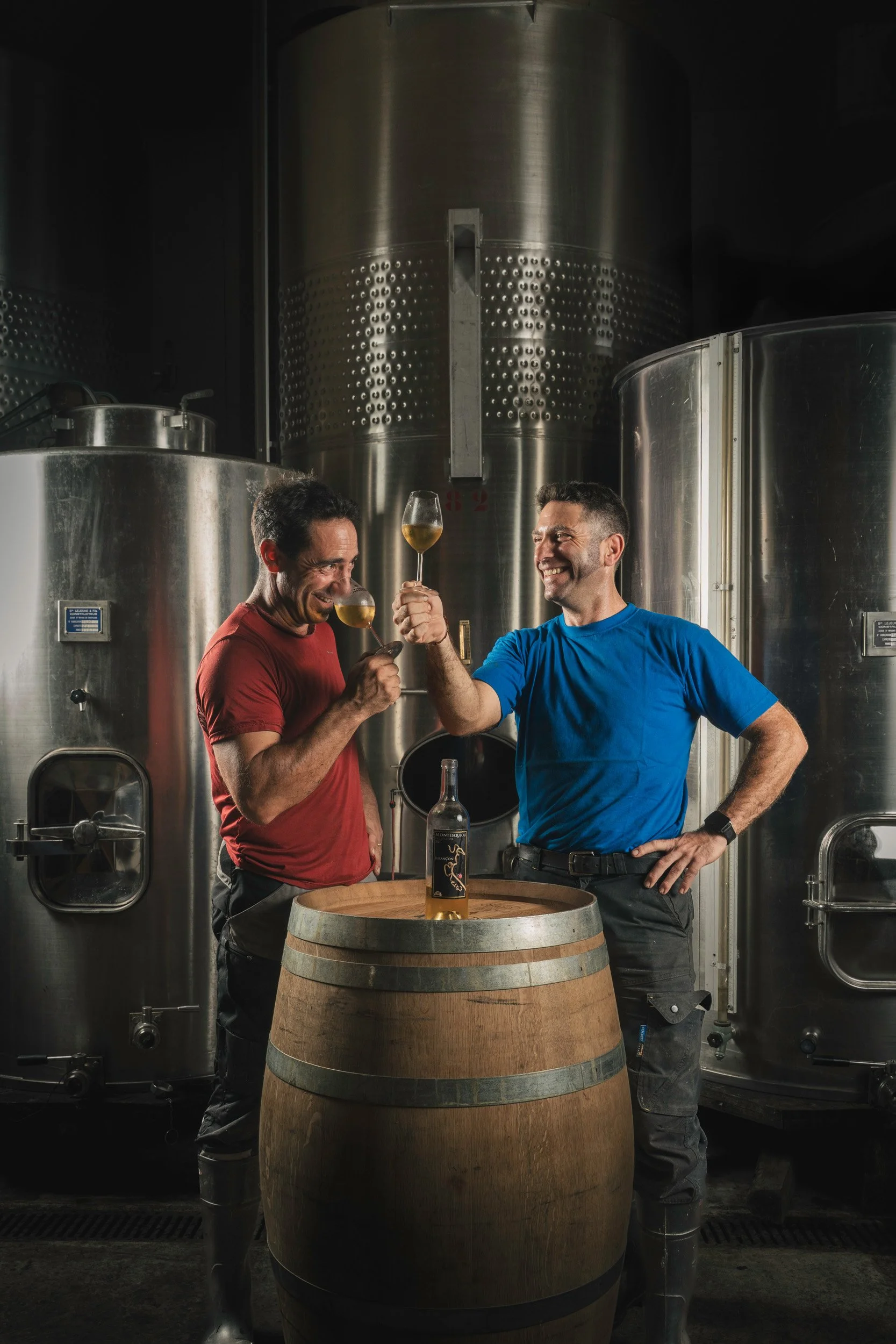 Two men in a brewery clinking glasses of wine or beer over a barrel, with large stainless steel brewing tanks in the background.