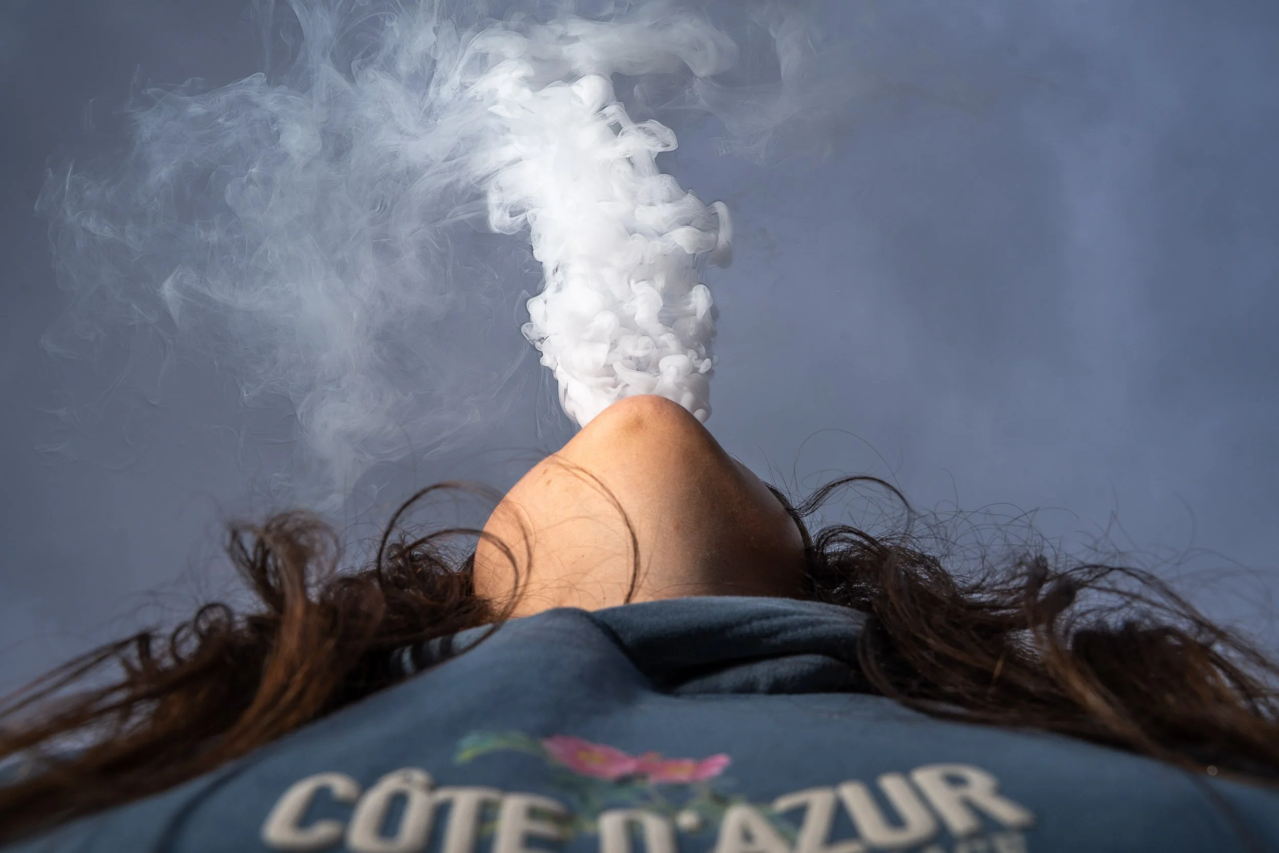 A person with long hair is standing outdoors, wearing a shirt with a floral design and the words 'Côte d'Azur' on it. The person is exhaling vapor or smoke, which is visible rising from their mouth.