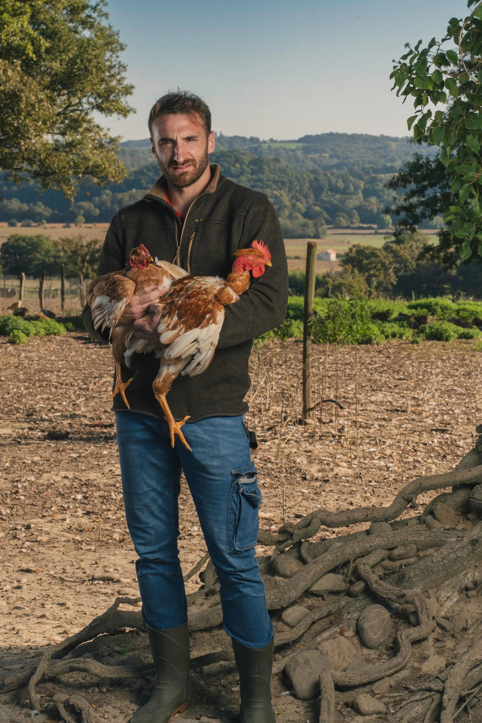 A man standing outdoors on a farm holding two chickens, with green fields and trees in the background.