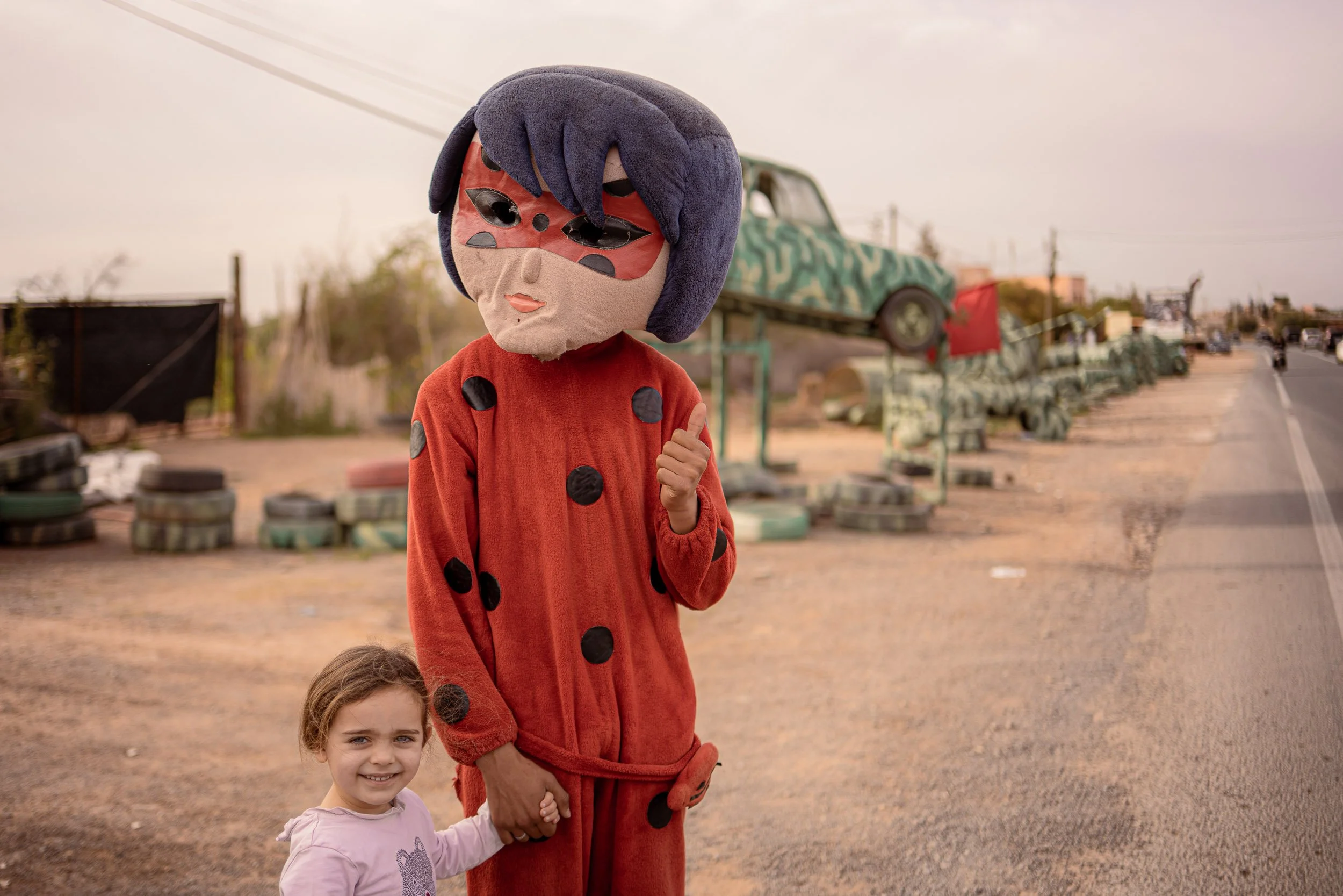 A person in a ladybug costume giving a thumbs up, holding a child's hand. The child is smiling, standing beside the costumed person. They are outdoors on a dirt road with an abandoned car and tires stacked in the background.