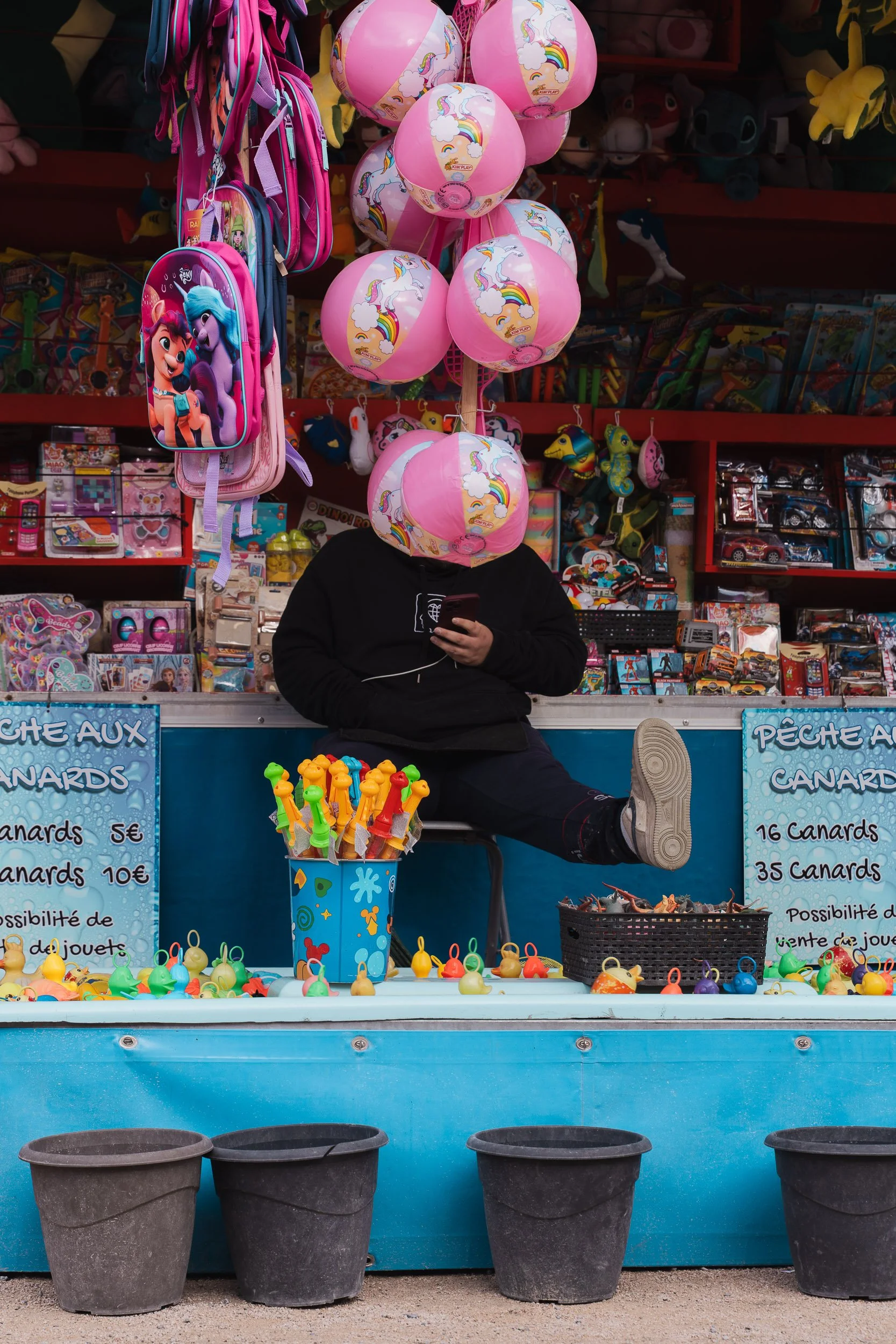 A street vendor stall selling rubber ducks, colorful balloons with rainbows and clouds, toy backpacks, and various toys. The vendor is sitting with their face hidden behind pink rainbow-patterned balloons, holding a phone, with legs crossed and resti
