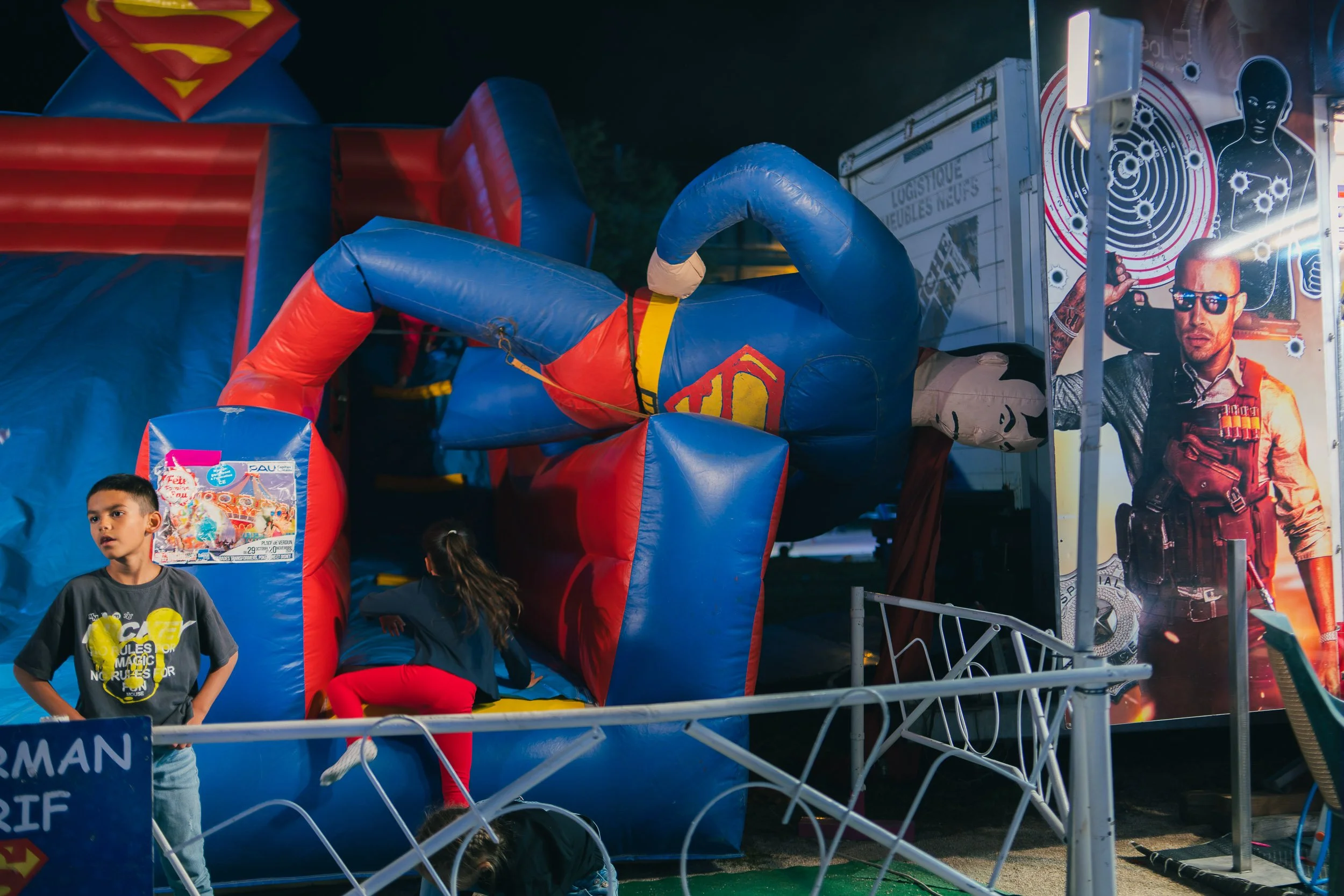 Children playing on an inflatable Superman-themed bounce house at night, with a promotional poster of a man in sunglasses on the right side.