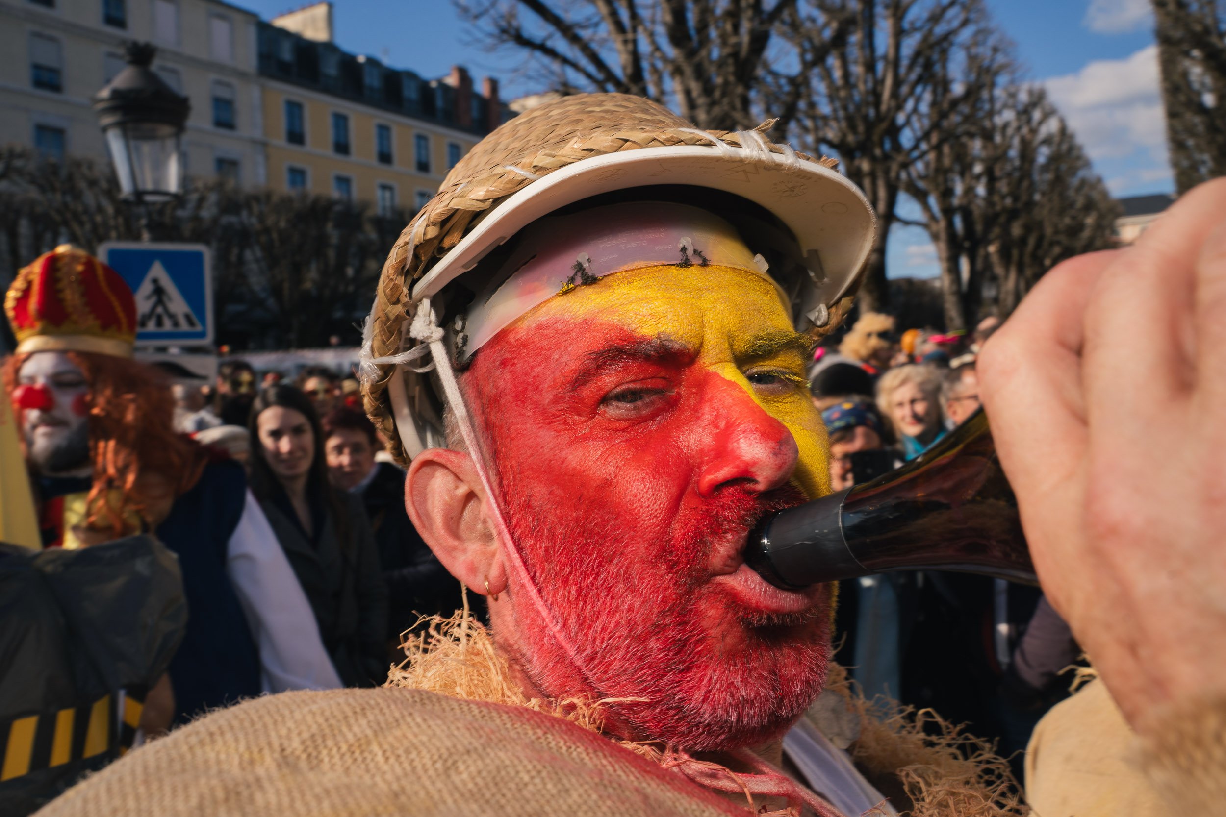 A person with face paint in red and yellow colors, wearing a straw hat and painting a helmet, is drinking from a black bottle in a crowd during a parade or celebration.