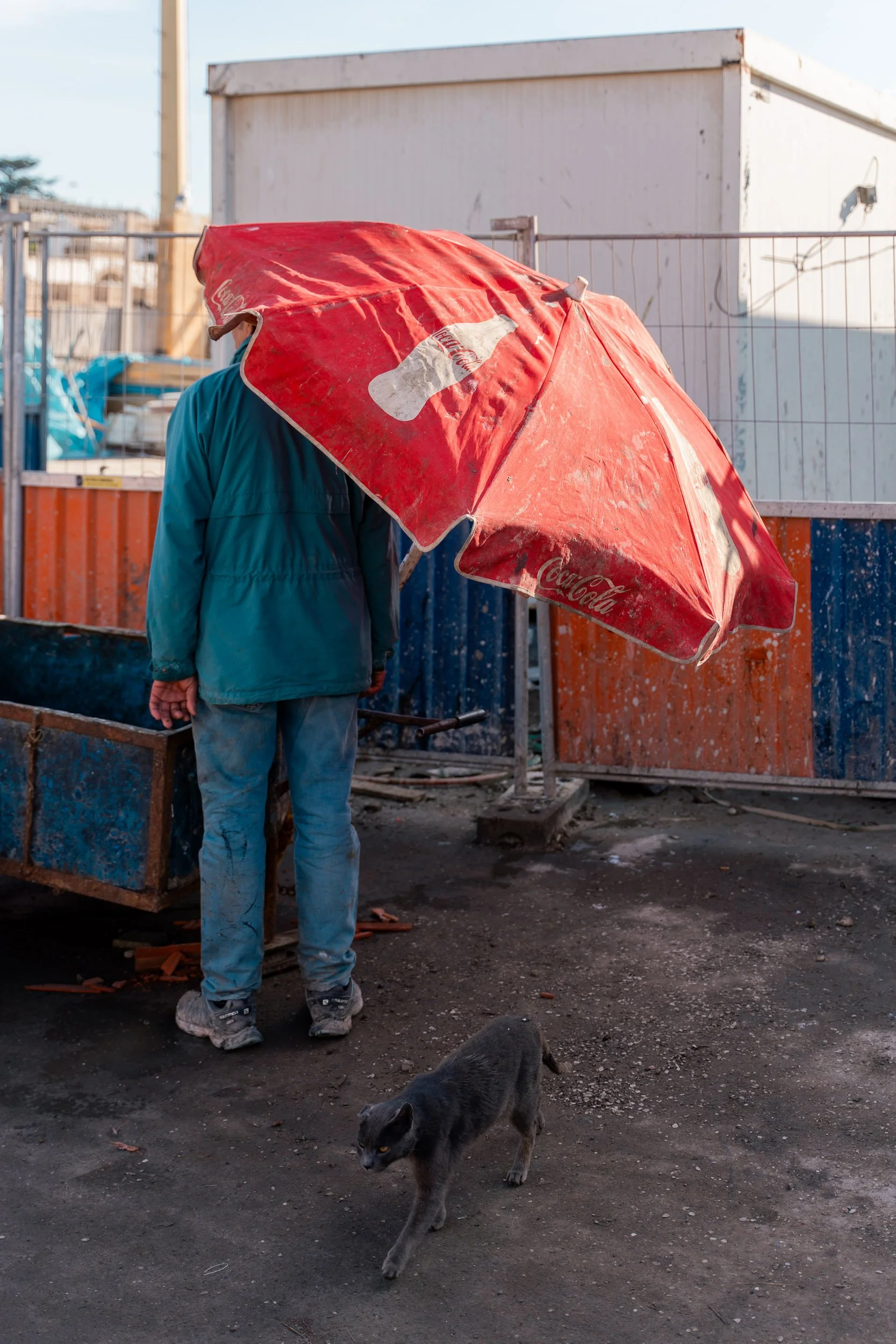 A person standing under a large red Coca-Cola umbrella in an outdoor setting, with a black cat walking on the ground nearby.