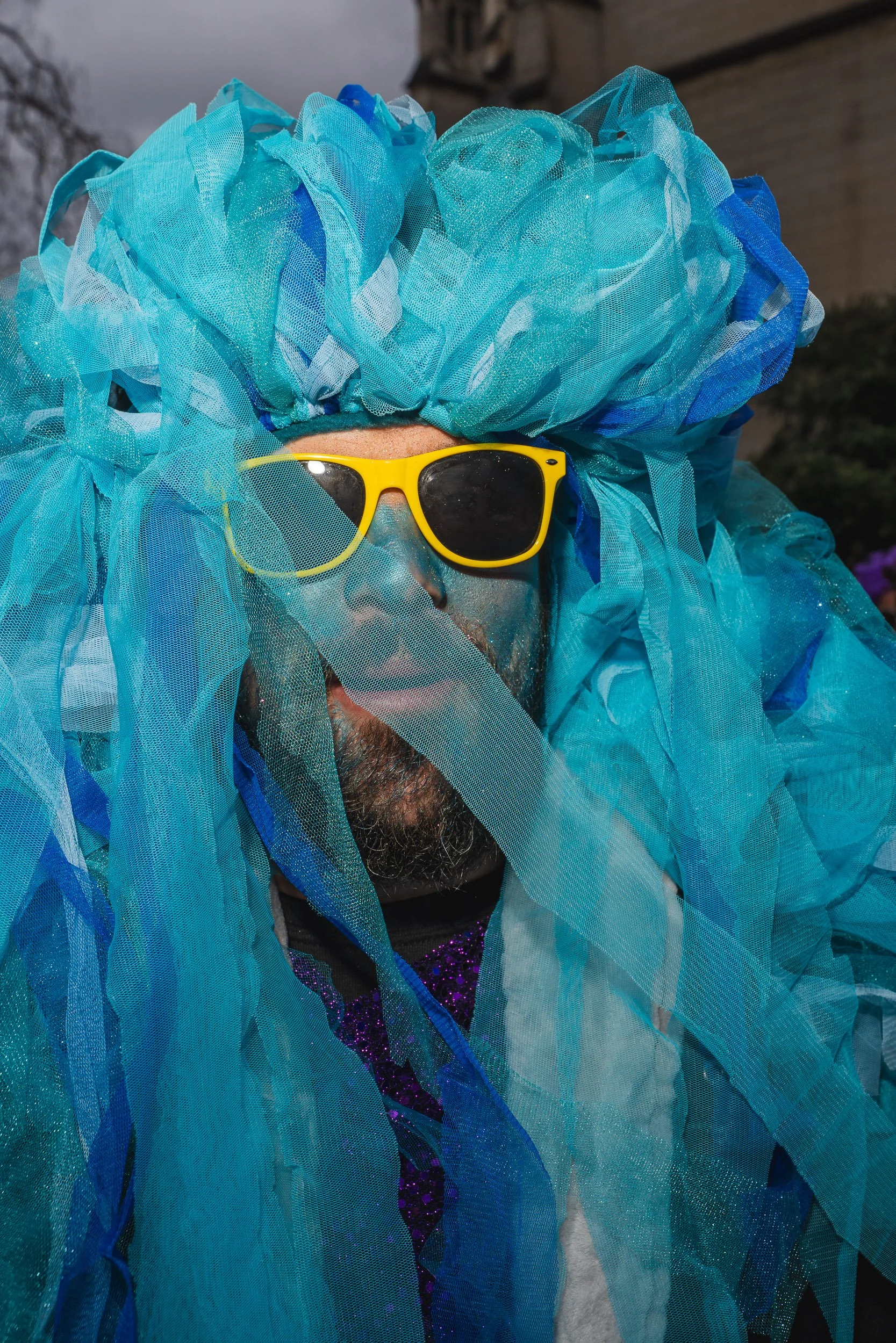 Person dressed in a vibrant blue costume with large, ruffled tulle, wearing yellow sunglasses, with face painted blue and glitter, outdoors during daytime.