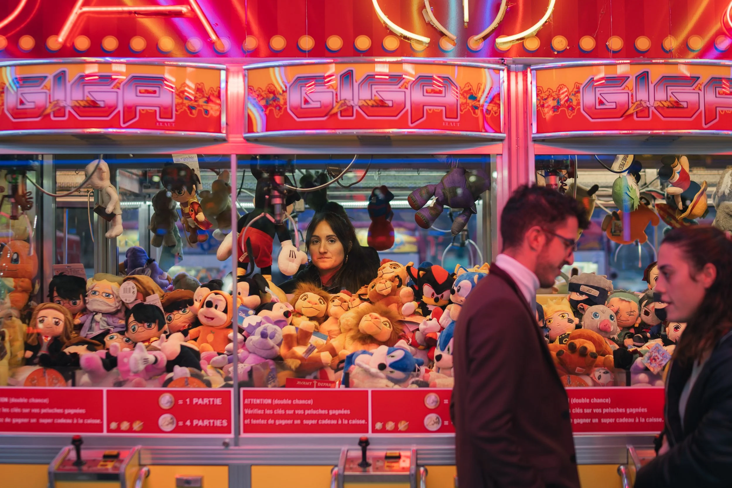 People at a carnival game booth filled with stuffed animal prizes, with a woman behind the counter and two people in the foreground talking, colorful neon lights