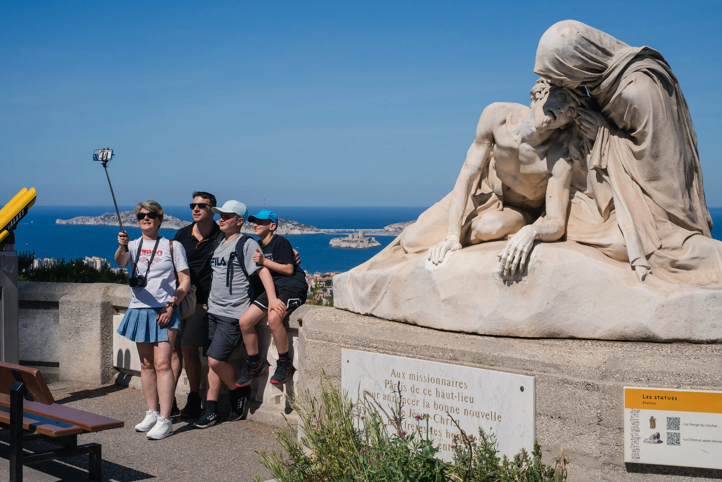 A family of four taking a selfie near a white marble statue of the Pietà, with a sea view and island in the background, on a sunny day.