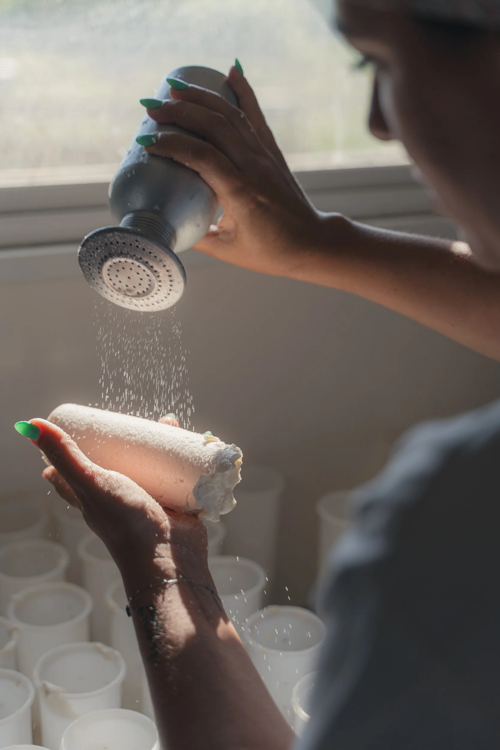 A woman with green nails spraying salt on a cone of local ice cream, standing near a window during daytime.