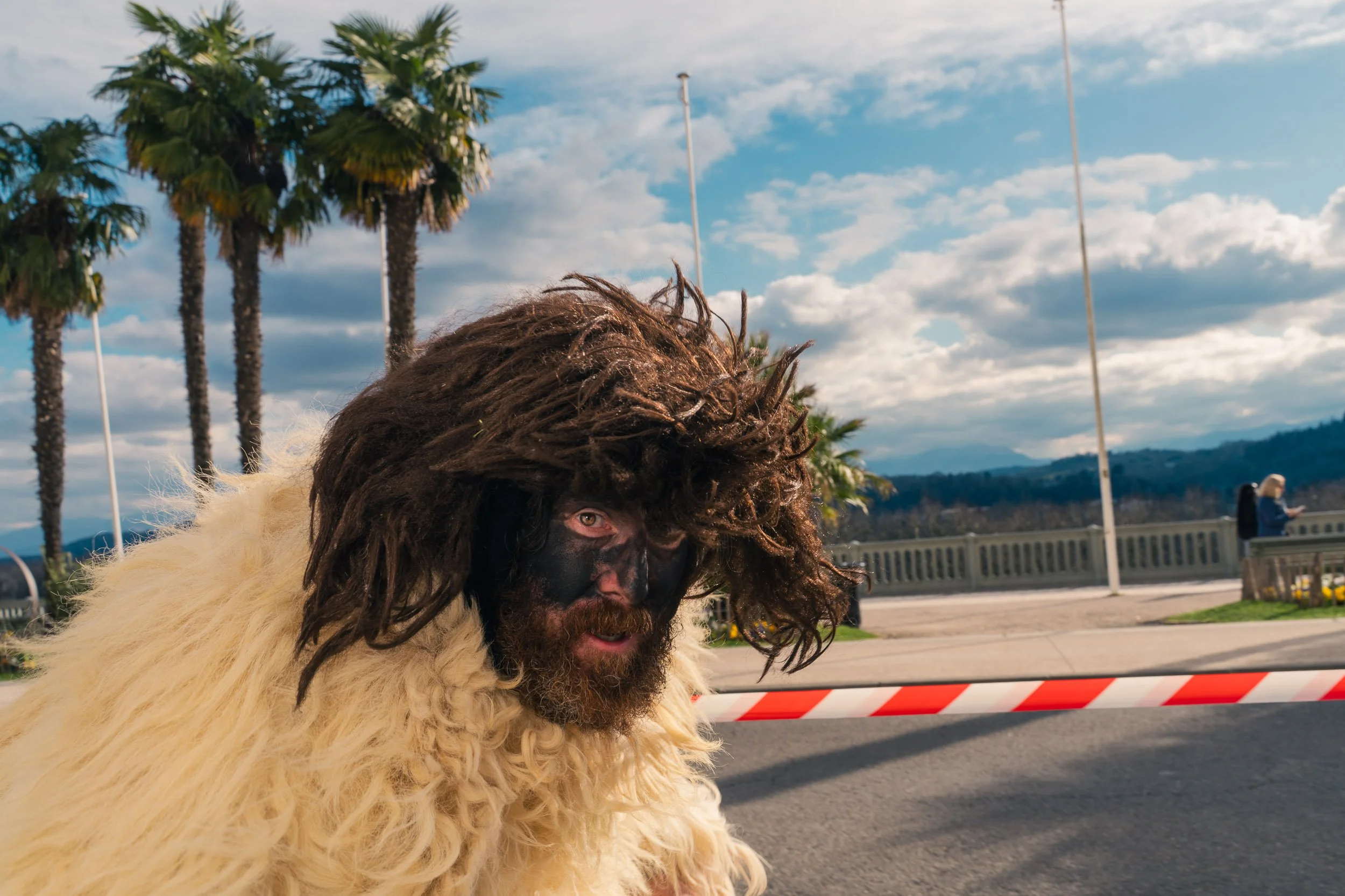 Person dressed in a costume resembling a yeti or sasquatch, with long dark hair, face paint, and a furry white outfit, standing outside near a barrier on a street with palm trees and a sky filled with clouds in the background.